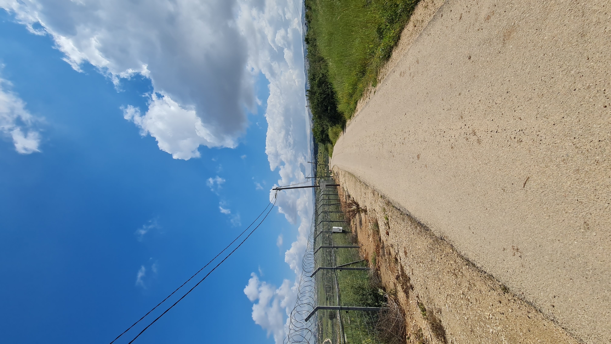 The image shows a rural scene in Hof Ashkelon Regional Council, Israel. A dirt road stretches into the distance, bordered on one side by green vegetation and on the other by a fence topped with barbed wire. Utility poles run parallel to the fence. The sky is blue with white, puffy clouds.