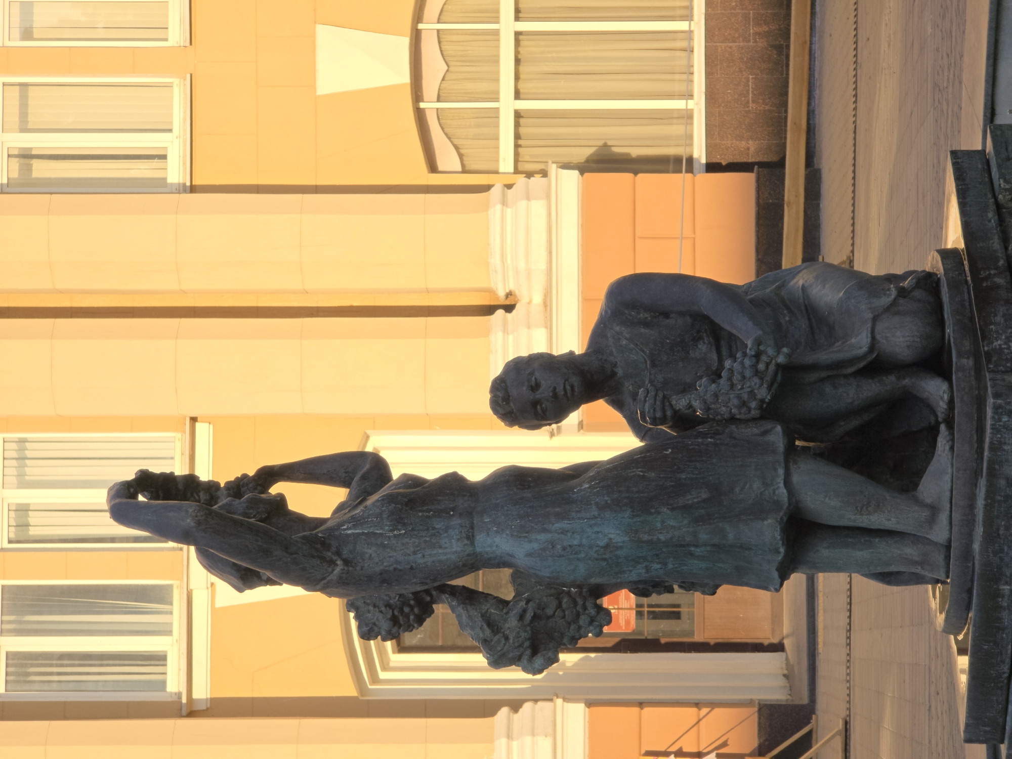 This is a vertical, eye-level, medium shot of a dark bronze sculpture of two young figures in front of a building with yellow walls and white columns. The sculpture depicts a standing female figure with grapes in her arms and a kneeling male figure also holding grapes. The standing figure's right arm is raised and her head is tilted back as if she is offering the grapes or celebrating. The kneeling figure's gaze is directed downwards, also towards the grapes. The sculpture is placed on a dark base in a paved outdoor area. The background building has tall, narrow windows with white frames and cream-colored curtains. The sunlight casts long shadows, suggesting it is late afternoon or early morning. This sculpture is located in Rostov-on-Don, Russia, in front of the Rostov-on-Don State Scientific Library.