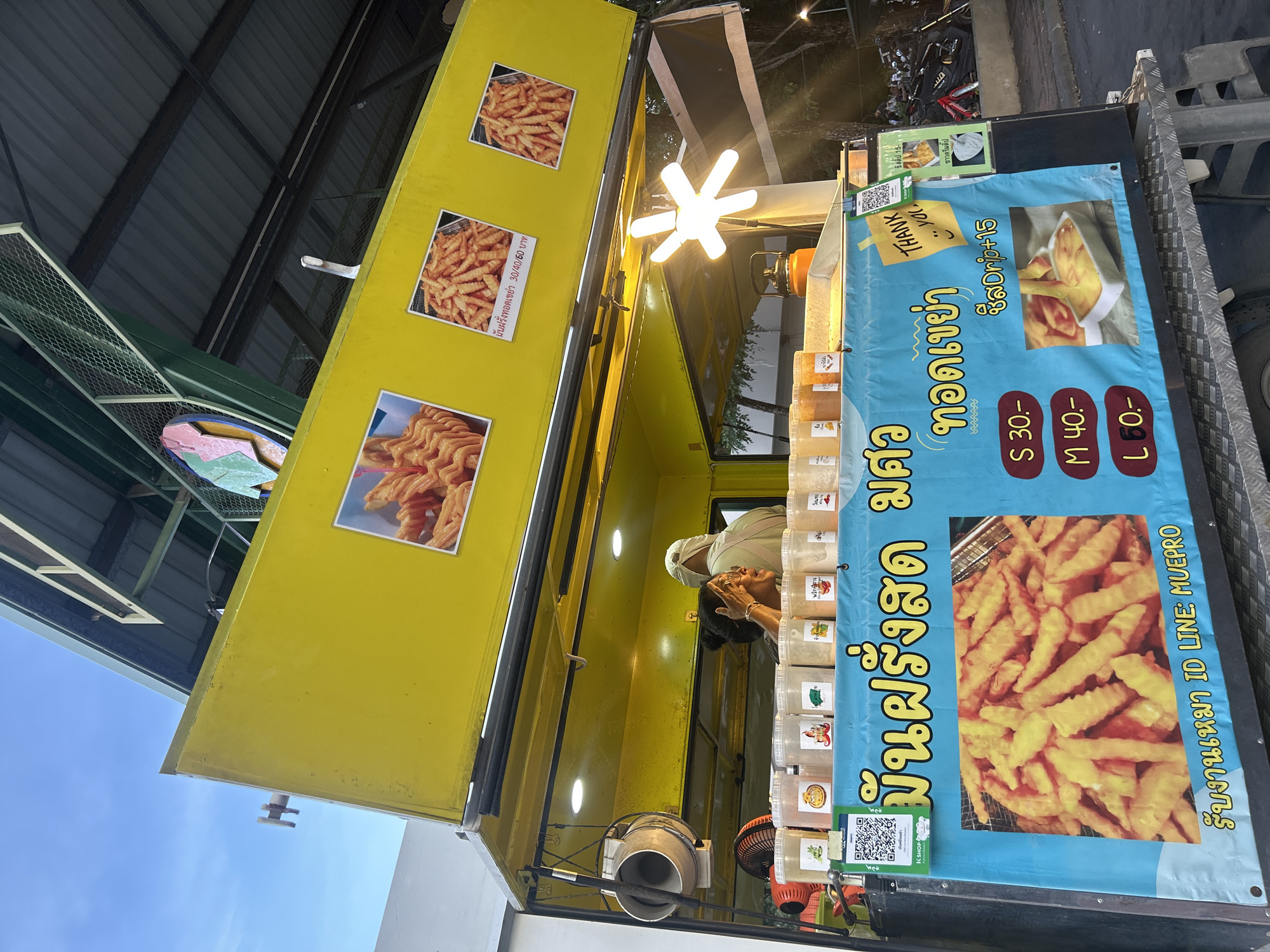 A food cart selling French fries is parked on a street in Tha Khlong Town Municipality, Thailand. The cart is yellow with a blue tarp displaying the menu. A woman is seen working at the cart, serving customers. The weather is overcast, and it appears to be late afternoon or early evening. The food cart is decorated with pictures of French fries and various dipping sauces. There are also QR codes for payment. The cart has a selection of seasonings displayed in clear containers. The street is paved, and there are trees and buildings visible in the background. A motorcycle is parked nearby, suggesting the area is a public space. The overall scene depicts a typical street food vendor operation in Thailand.