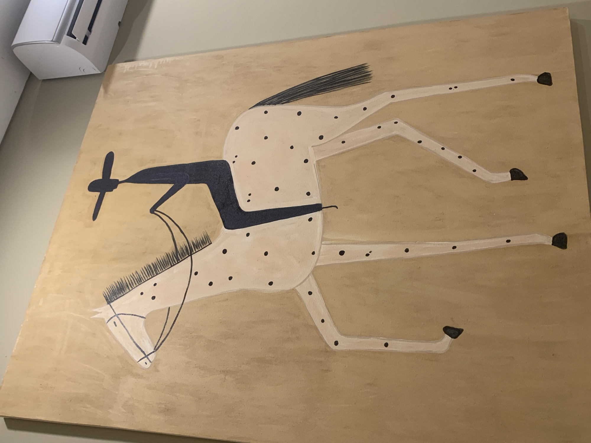 This media file is a close-up, angled shot of a painting on a light brown, possibly wood, surface. The painting depicts a stylized, white horse with black polka dots. The horse is shown in profile, with its body elongated and its legs splayed out in a dynamic, almost airborne pose. Its mane and tail are rendered as short, dark brushstrokes.
The horse has a dark blue saddle-like shape on its back. Attached to its head is a dark blue, propeller-like object with a central circle and two crossbars, suggesting a whimsical or fantastical element. The overall style is simplistic and somewhat abstract, with thin black lines outlining the horse's head and neck.
The background of the painting is a uniform light brown, providing a neutral canvas for the artwork. In the top left corner of the image, a portion of a white object is visible, possibly an air conditioning unit, casting a shadow on the wall behind it.
The painting does not appear to depict a specific event or location, but rather a singular, imaginative subject. There is no visible text in the image. The lighting suggests it is indoors, possibly during the day or under artificial light, and there are no cues about weather or a specific time of day. The emotion conveyed by the artwork is one of movement and fantasy. The location context of North Kuta, Indonesia, does not seem to be visually represented in the painting itself.