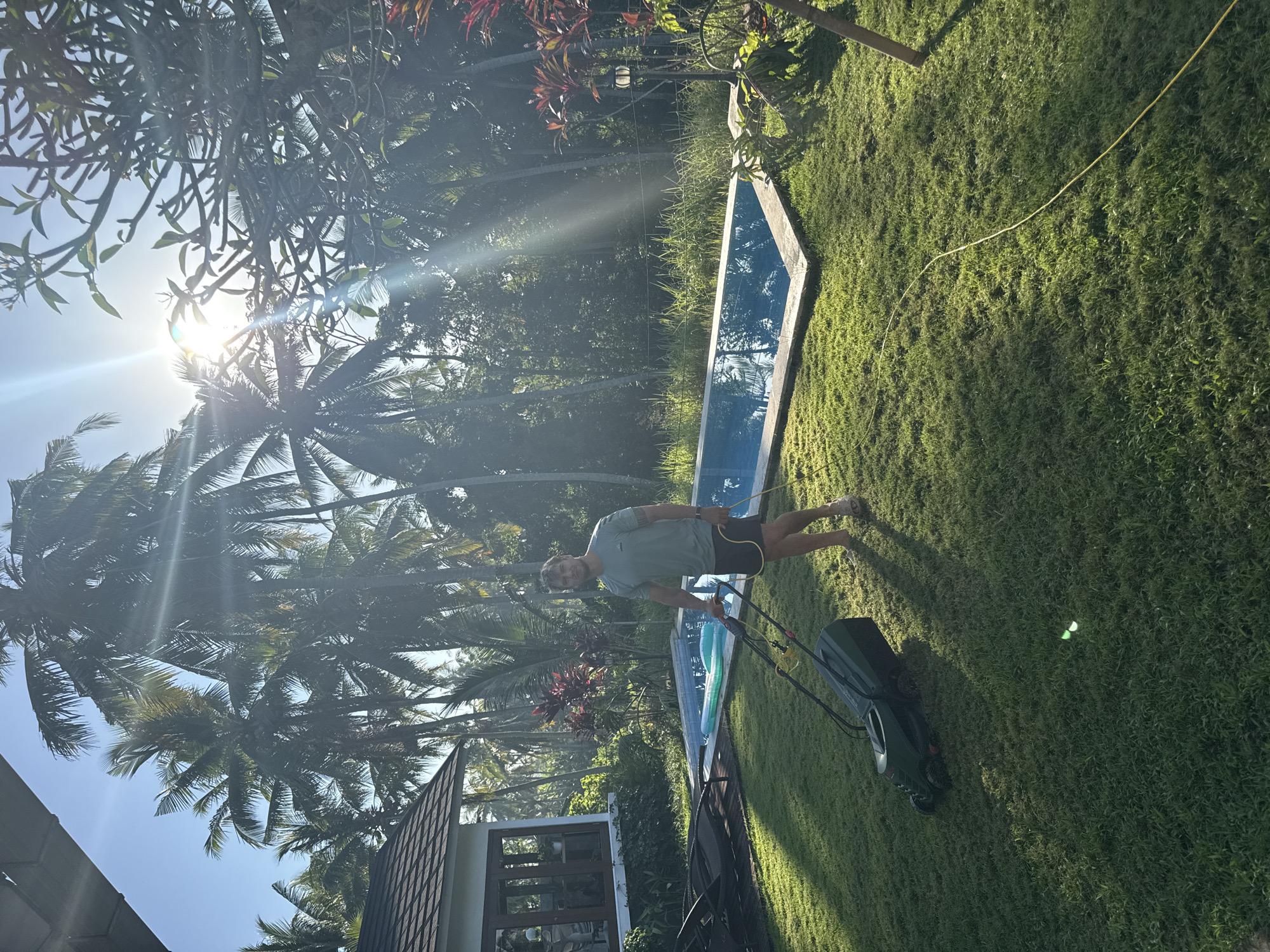 A man is mowing a grassy lawn in what appears to be a tropical location in Unknown, Indonesia. He is wearing a light-colored t-shirt and dark shorts. He is holding a green and black electric lawnmower. A yellow extension cord is visible on the lawn. Behind him, there is a rectangular pool with blue water. Tropical foliage, including palm trees, is visible. There is also a house with a dark roof in the lower left corner of the image. The sun is shining brightly through the trees.
