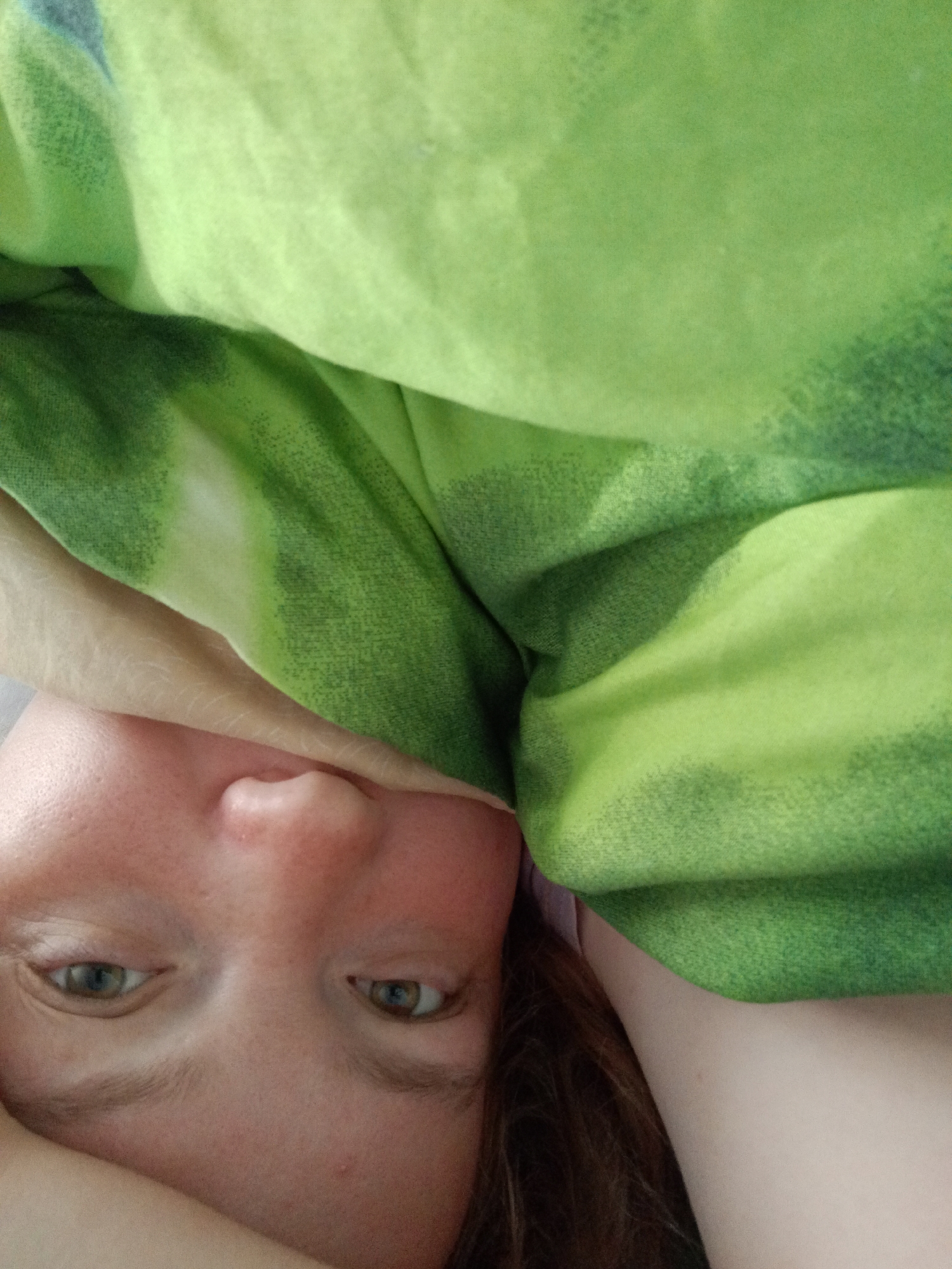 The image shows a person's face and part of their upper body under a green and light green patterned blanket. The person has fair skin, light brown hair, and green eyes. The image is taken from a close-up, slightly rotated perspective. No location or city can be discerned from the image.