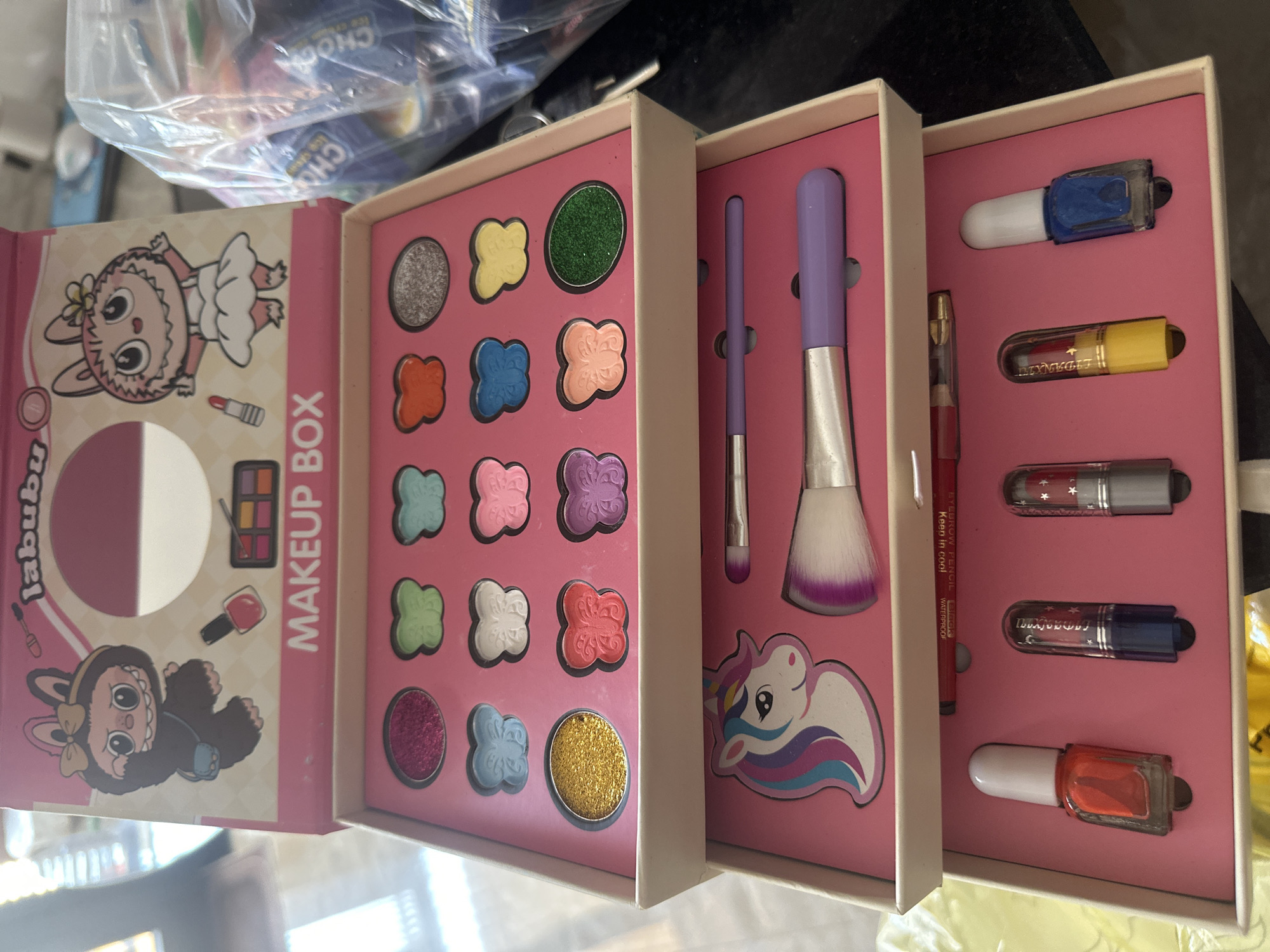Here is a detailed description of the image:
**Scene:** An eye-level, close-up shot of a children's makeup box set.
**Objects:** The makeup set is pink and white. The open box reveals several compartments. The first compartment contains eyeshadows in various colors (silver, yellow, green, orange, blue, peach, teal, pink, purple, light green, white, coral, magenta, baby blue, and gold). The second compartment contains makeup brushes and a sticker of a unicorn. The third compartment contains various small makeup items, including what appear to be nail polishes in red, yellow, white, and blue.
**Environment:** The background appears to be a cluttered domestic setting with various other objects out of focus. There is no indication of the scene taking place in the city of Bac Nha Trang Ward, Vietnam.