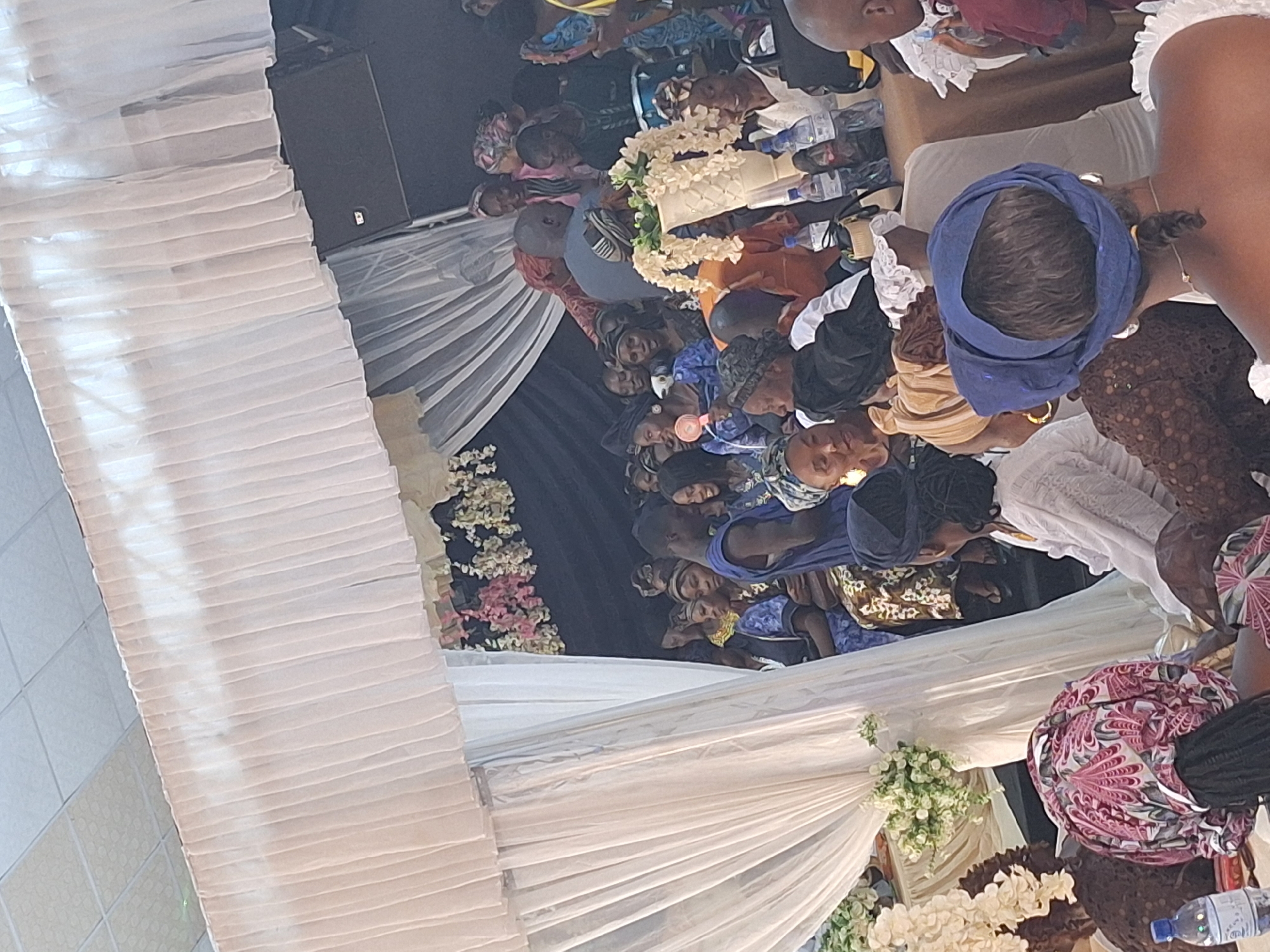 A slightly rotated high-angle shot shows a large group of people seated, primarily women, in a decorated event space, likely in Sabon Tasha, Nigeria. The event space has draped white fabric and flower arrangements of white and pink flowers. Many people are wearing head wraps and traditional clothing. There are bottled water and other items on tables in front of the seated guests. A speaker is set up in the background. The location appears to be a banquet hall or a similar venue set up for a gathering.