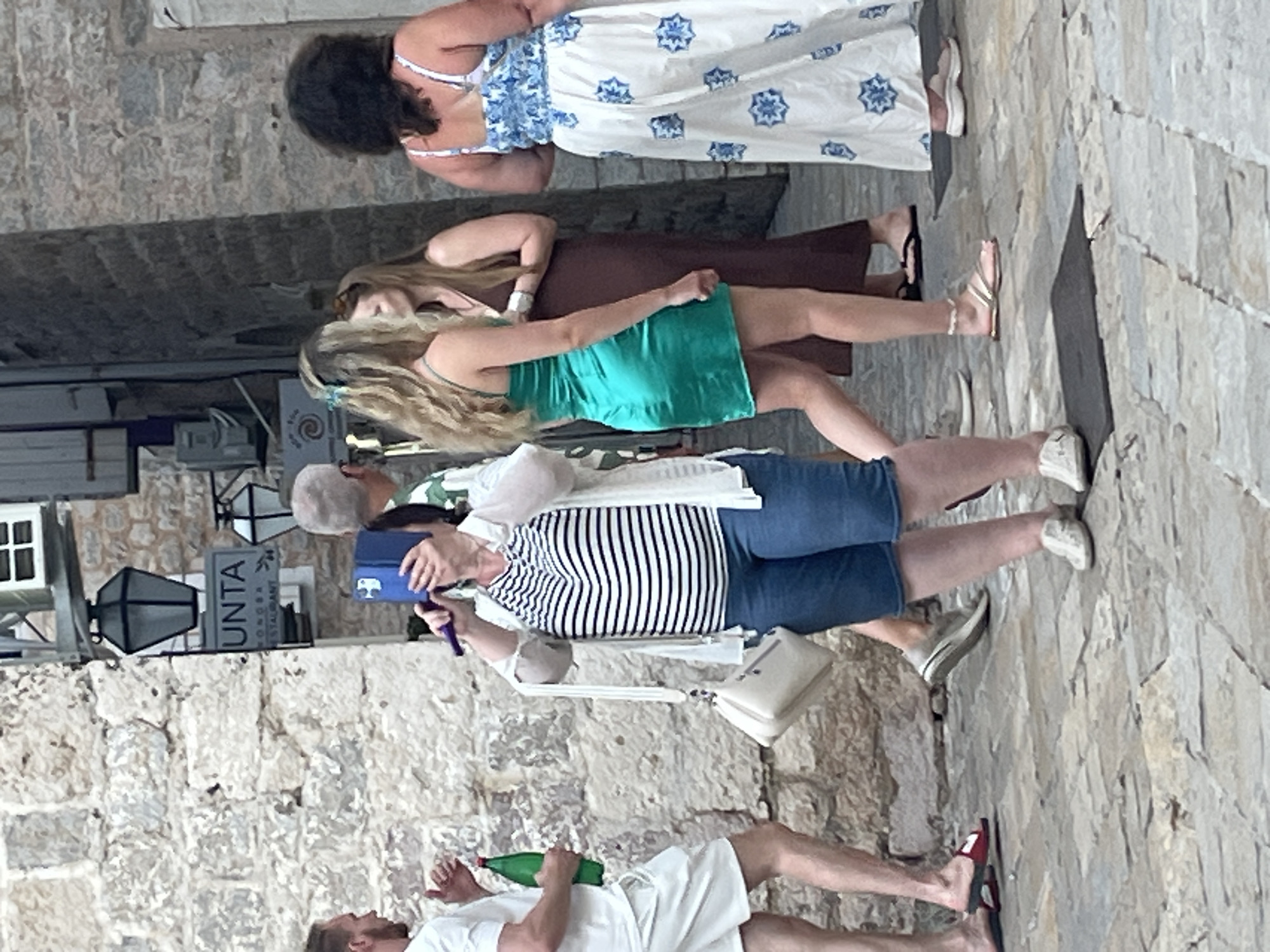 The image shows a street scene in Budva, Montenegro, with a stone building on the left and a cobblestone street in the foreground. Several people are walking on the street. On the left, a man in white shorts and a t-shirt holds a green bottle. In the center, a woman in a striped shirt and blue shorts is holding a blue object to her face. To her right, a woman in a green dress walks past, followed by a woman in a dark dress and another woman in a white dress with a blue floral pattern. The street is paved with large, irregular stones, and there is a stone wall on the left side of the image. A sign with the word "UNTA" is visible on the building.