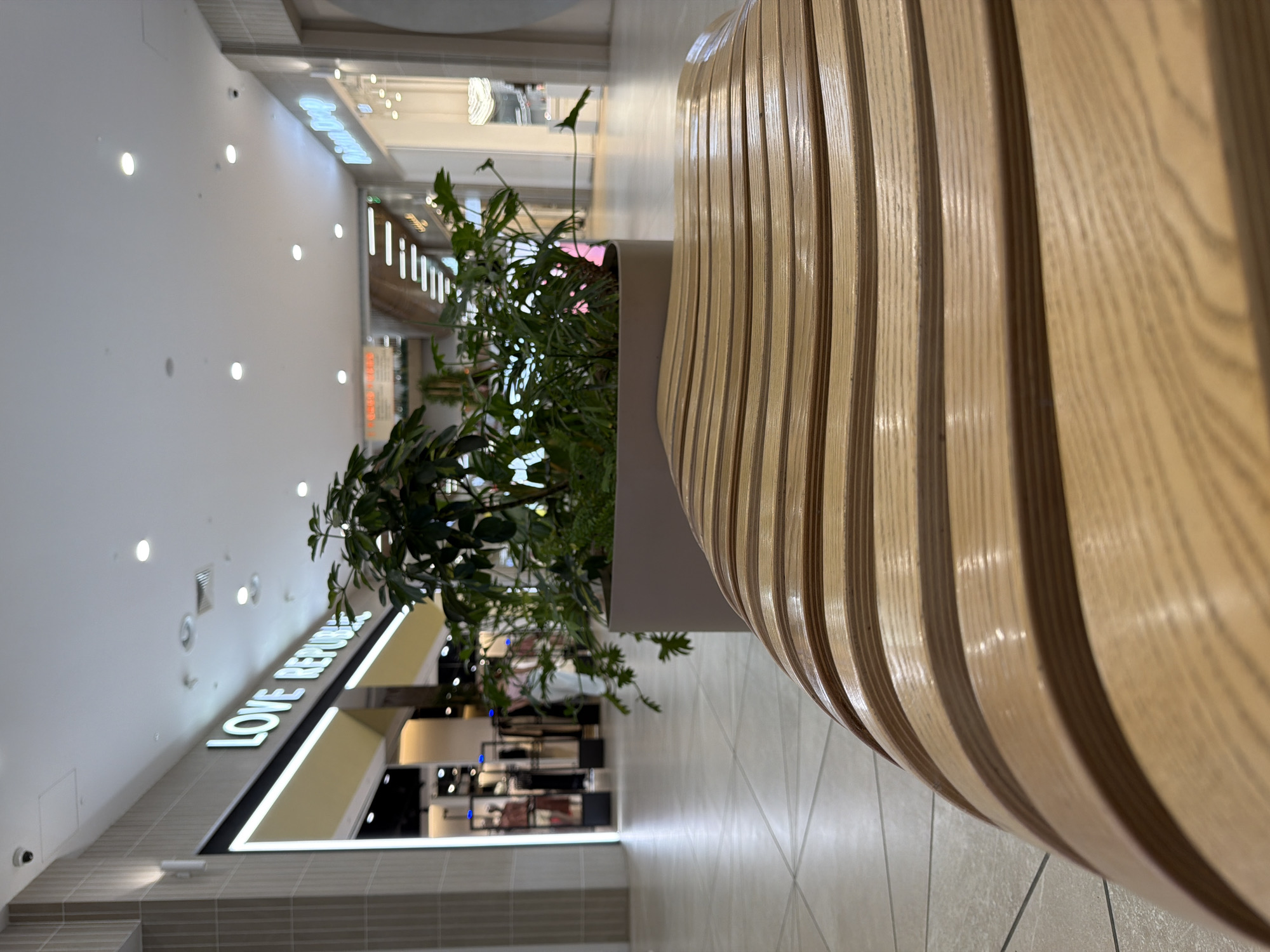 The image depicts an interior scene, likely within a shopping mall in Kazan, Russia. The foreground is dominated by a curved wooden structure with a slatted design, possibly a bench or decorative element. A planter containing greenery is positioned adjacent to this structure.
In the background, a long corridor stretches into the distance, lined with retail stores. The store "LOVE REPUBLIK" is visible on the left, with illuminated signage. The lighting is bright and modern, illuminating the clean, tiled floor. There are no people visible in the image, suggesting a quiet moment or a focus on the architectural and design elements. The overall environment suggests a contemporary and well-maintained commercial space.