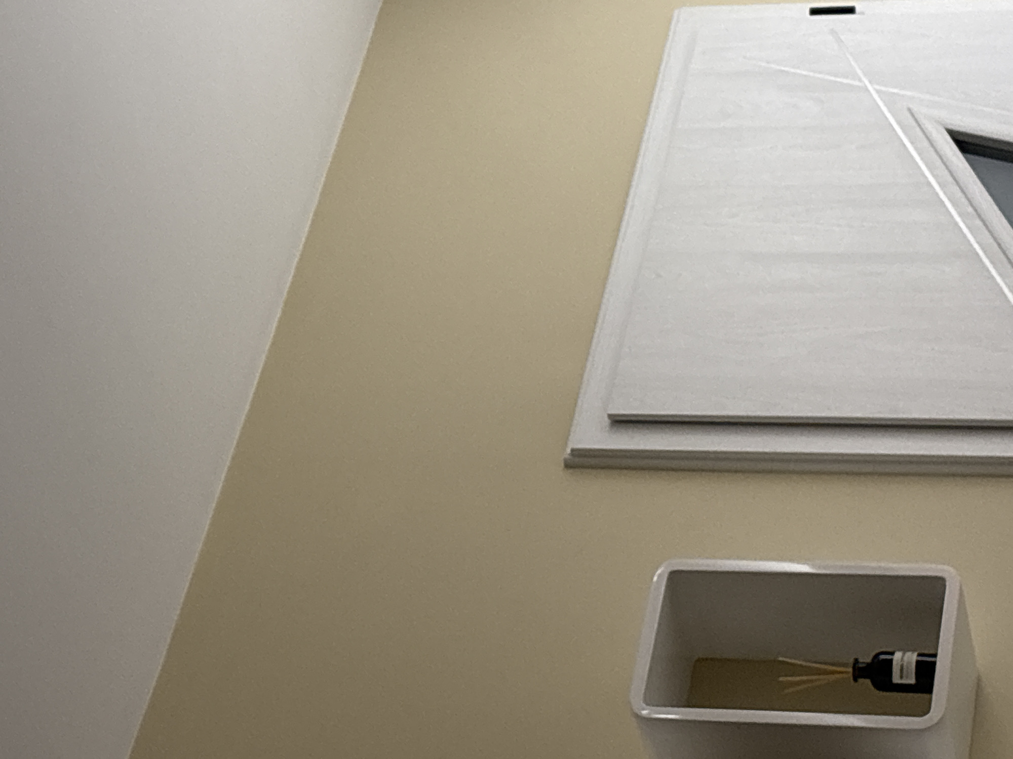 The image displays an interior scene, likely within a building in Sofia, Bulgaria. The view is angled upwards, showcasing a corner where two walls meet, along with elements attached to one of the walls.
On the left, a plain, off-white or very light gray wall occupies the majority of that side of the frame. It meets a light beige or tan-colored wall that extends across the center and right side of the image. Both walls have a smooth, matte finish.
On the beige wall, towards the upper right, there is a large, light-colored panel, possibly a door or a decorative wall feature. This panel is white or very light gray with a visible texture, possibly resembling wood grain. It features several raised, linear designs, including a prominent diagonal line creating a triangular shape in its lower right section. A small, dark, rectangular recess or slot is visible along the top edge of this panel.
In the lower right portion of the beige wall, a white, rectangular, wall-mounted or recessed holder is visible. Inside this holder, a dark glass bottle, resembling a reed diffuser, is placed. Several slender, light-colored reeds extend upwards from the bottle. A white band or label is wrapped around the bottle, but no text is legible.
The scene is static, with no people or activities depicted. The lighting suggests an indoor setting, with no indications of the time of day or outdoor weather. There are no specific location cues within the image itself beyond the provided context of Sofia, Bulgaria.