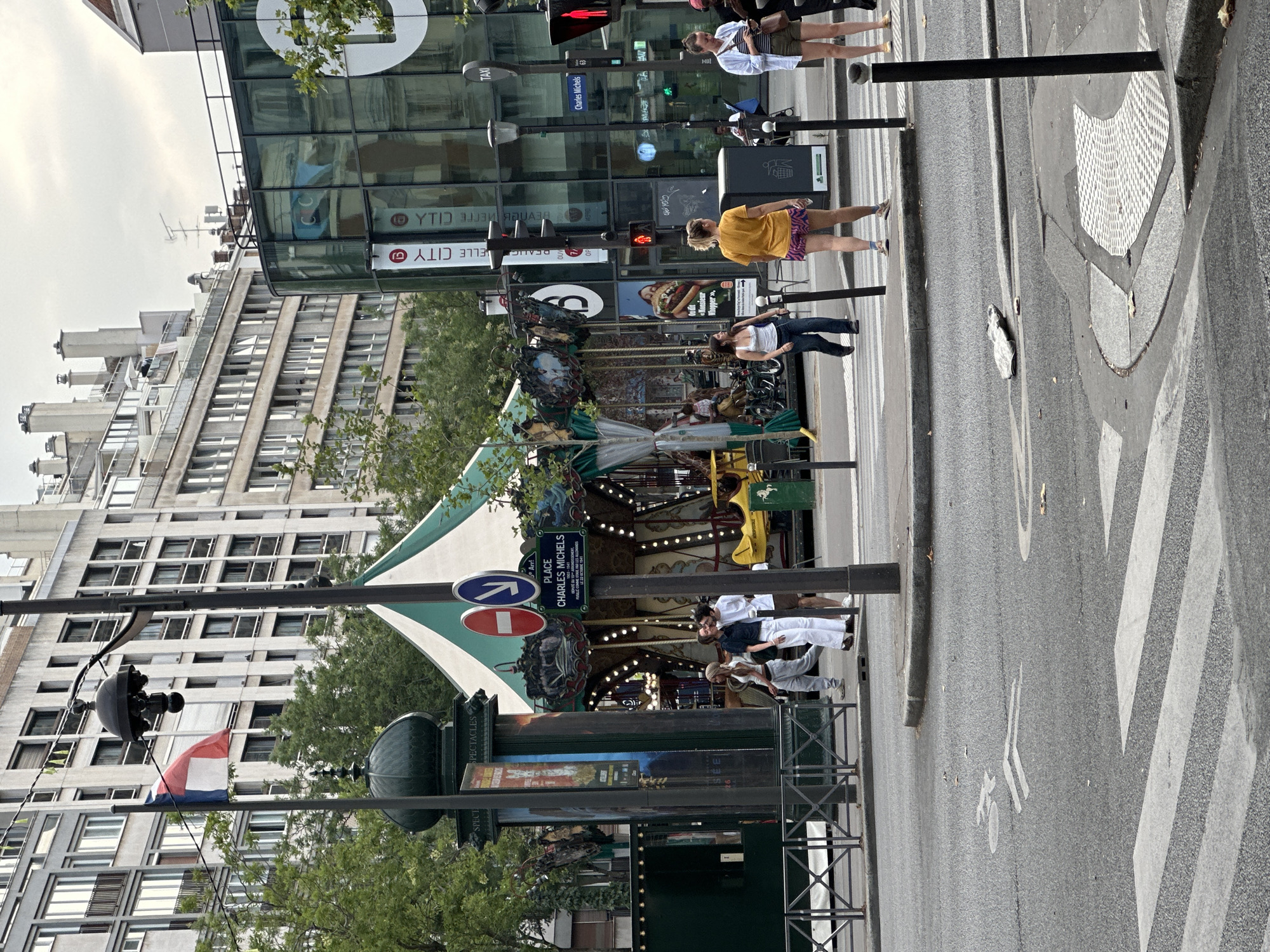 The image depicts a street scene in Paris, France, specifically at Place Charles Michels. In the foreground, a pedestrian crossing is visible on a paved road, with a bicycle lane marking painted on the asphalt. A manhole cover with a textured surface is also present. Beyond the road, a carousel with a green and white striped canopy is set up. Several people are seen walking and standing near the carousel and on the sidewalk. One man and two women are gathered near a lamppost in front of the carousel, appearing to be in conversation. Further to the right, two women are walking, one in a yellow shirt and shorts, and another in a white shirt and shorts, looking at their phones. In the background, tall, multi-story buildings with numerous windows line the street. A French flag is flying from a flagpole attached to a lamppost. The sky is overcast.