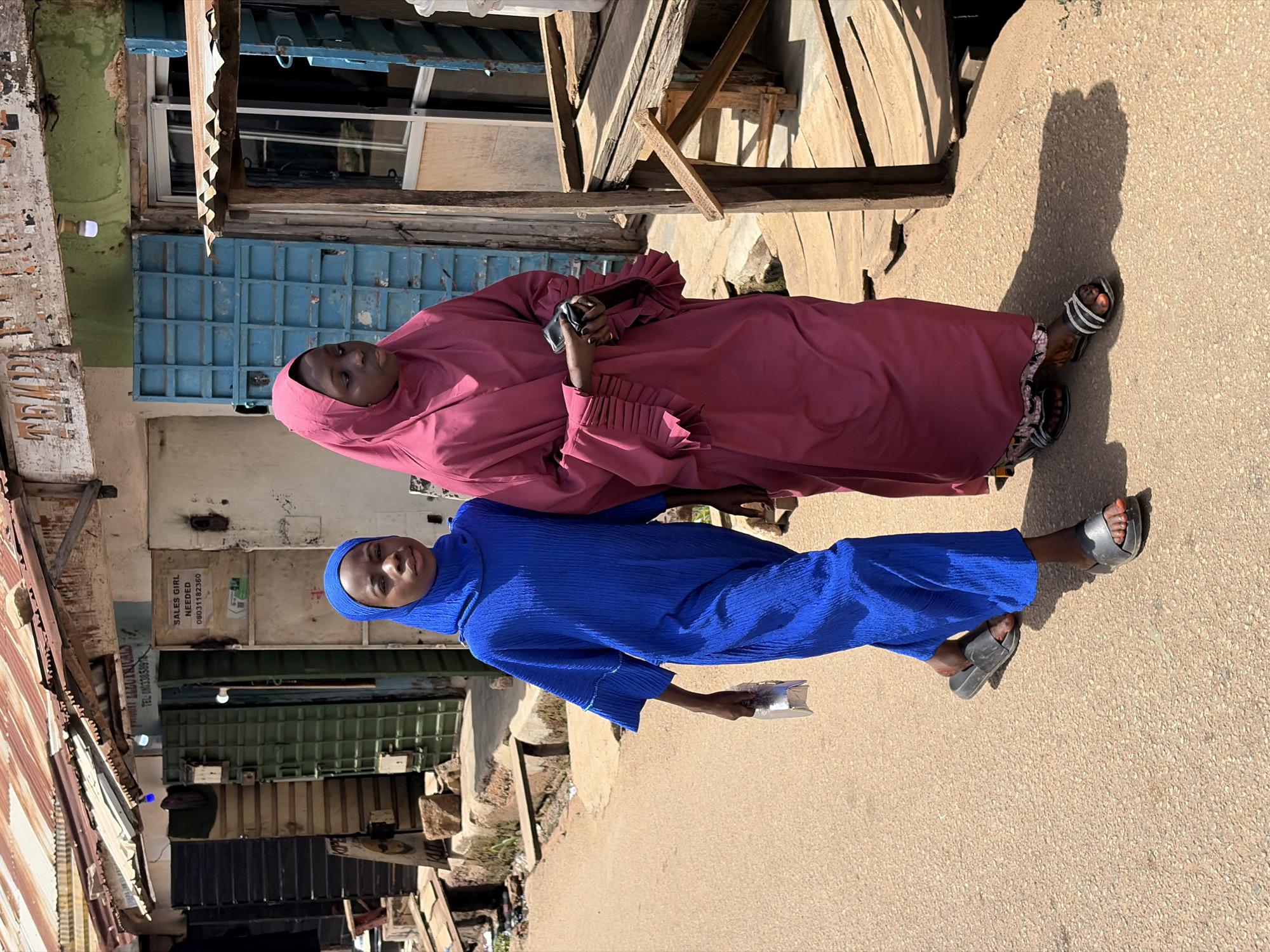 Two women stand on a paved surface beside a street in what appears to be the city of Jos, Nigeria. The woman on top wears a pink hijab and matching robe, holding what looks like a cell phone. The woman below wears a blue hijab and a matching outfit. The street is lined with buildings, including structures with corrugated metal roofs. Various signs are visible, including one that states, "SALES GIRL NEEDED." The background shows a mix of building materials.