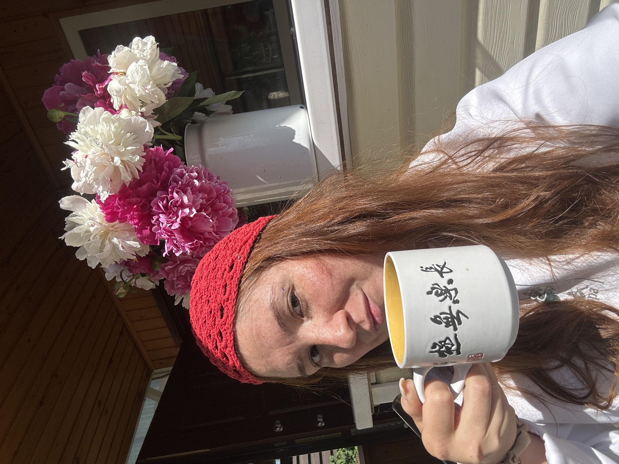 A woman with long, auburn hair and a red knitted headscarf holds a white mug with yellow interior and East Asian characters. She is wearing a white hooded sweatshirt. Behind her, a bouquet of pink and white peonies is in a white pitcher. The scene appears to be outdoors, possibly on a porch or patio with wooden siding and a wooden ceiling. The lighting suggests it is daytime, with strong sunlight creating shadows. The background includes a glimpse of a window and vegetation. The woman is looking towards the camera, and her right hand is holding the mug.