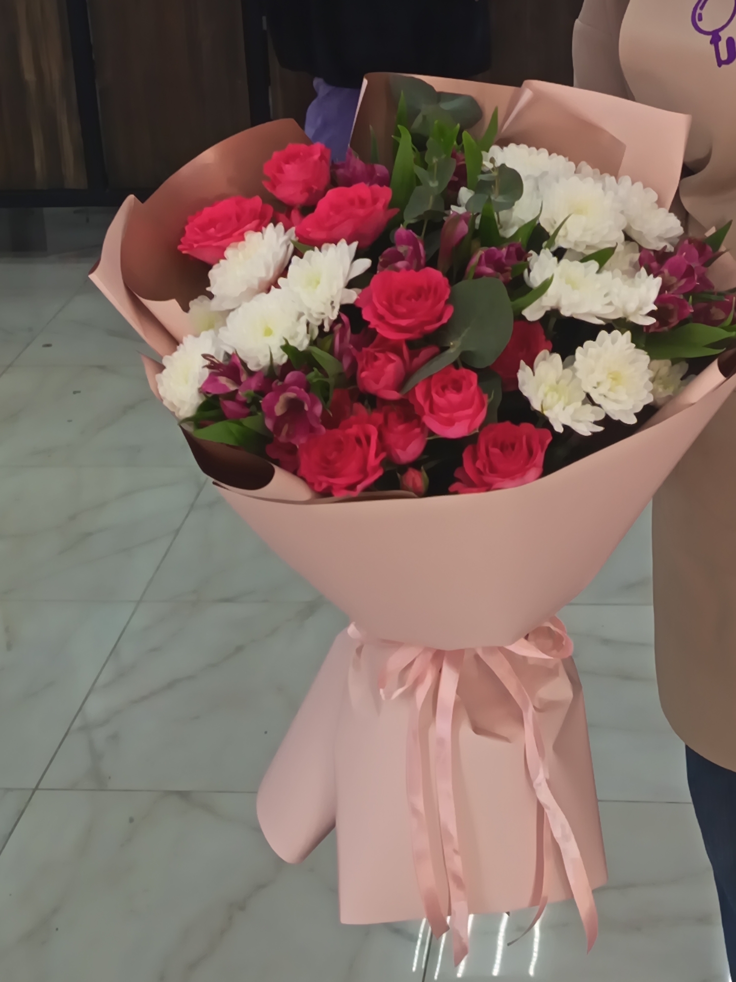 A large, colorful bouquet of flowers is prominently displayed, held by a person partially visible on the right side of the image. The bouquet features a rich assortment of red roses, fluffy white chrysanthemums, and vibrant purple alstroemeria, complemented by green eucalyptus leaves or similar foliage. It is elaborately wrapped in layered light pink and kraft-colored paper, secured at the base with a delicate pink satin ribbon tied into a bow.
The person holding the bouquet is seen from the chest down, wearing a light brown or tan long-sleeved top and dark blue trousers. A small, blurry purple graphic, resembling a stylized magnifying glass or abstract logo, is visible on the upper right side of their top.
The setting appears to be indoors, with a light-colored, possibly marble, tiled floor occupying the lower left and central foreground. The tiles are a light grey or white with darker veining. In the upper left background, a dark, indistinct wall or piece of furniture is visible. The overall impression suggests a celebratory moment or the act of giving a gift in Nizhnevartovsk, Russia. The lighting is consistent with an indoor environment.
