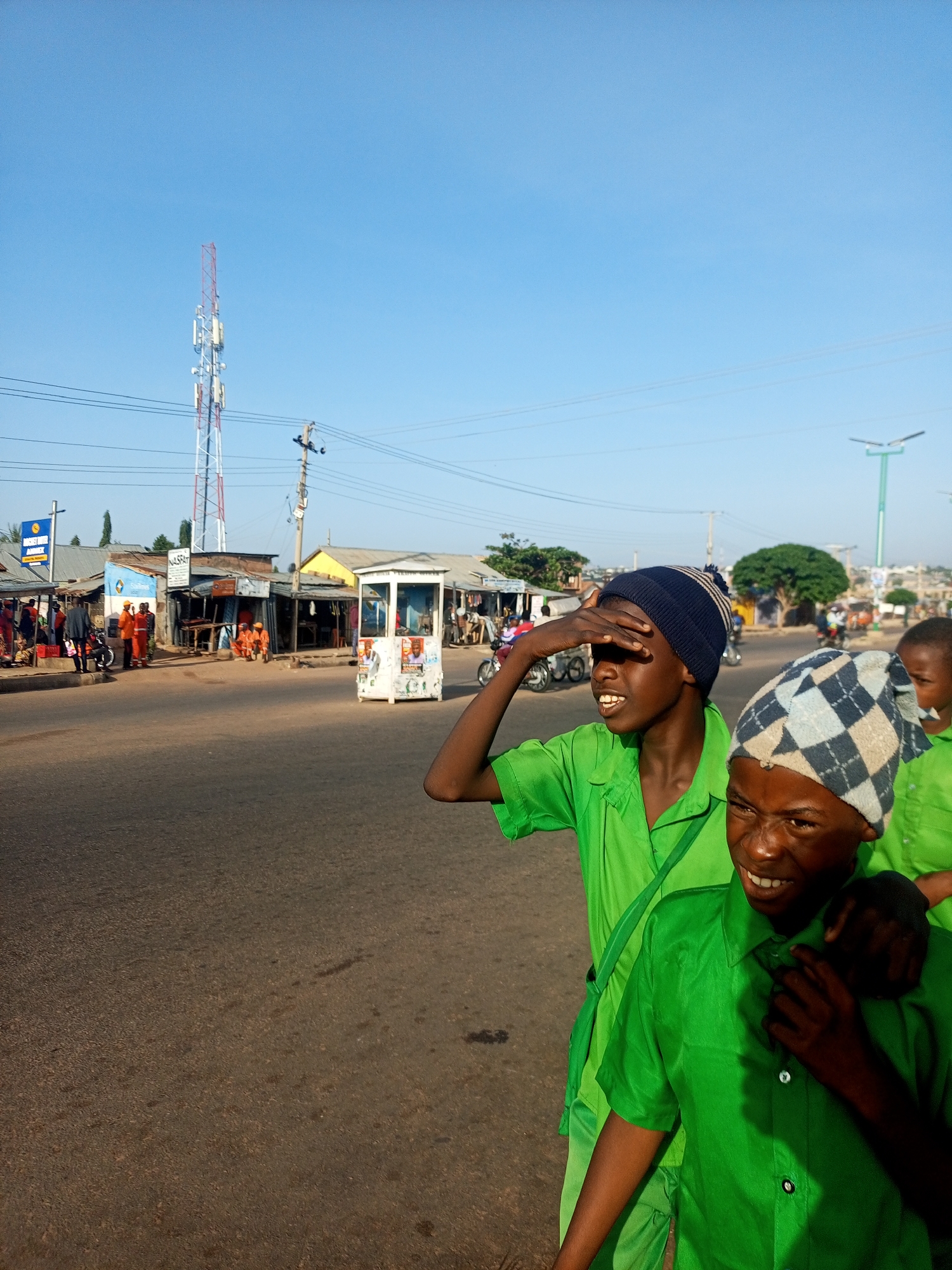 In the city of Unknown, Unknown, a street scene unfolds under a clear blue sky. In the foreground, two young boys, dressed in bright green shirts and hats, are the primary subjects. One boy, on the left, shields his eyes with his hand, gazing into the distance. The other boy, on the right, smiles broadly, looking towards the viewer or a point just off-camera. Behind them, the street is lined with small shops and market stalls, a telecommunications tower stands tall in the background, and power lines crisscross the sky. Several motorcycles are visible, suggesting transportation and activity in the area. The overall impression is a snapshot of everyday life in a bustling urban environment.