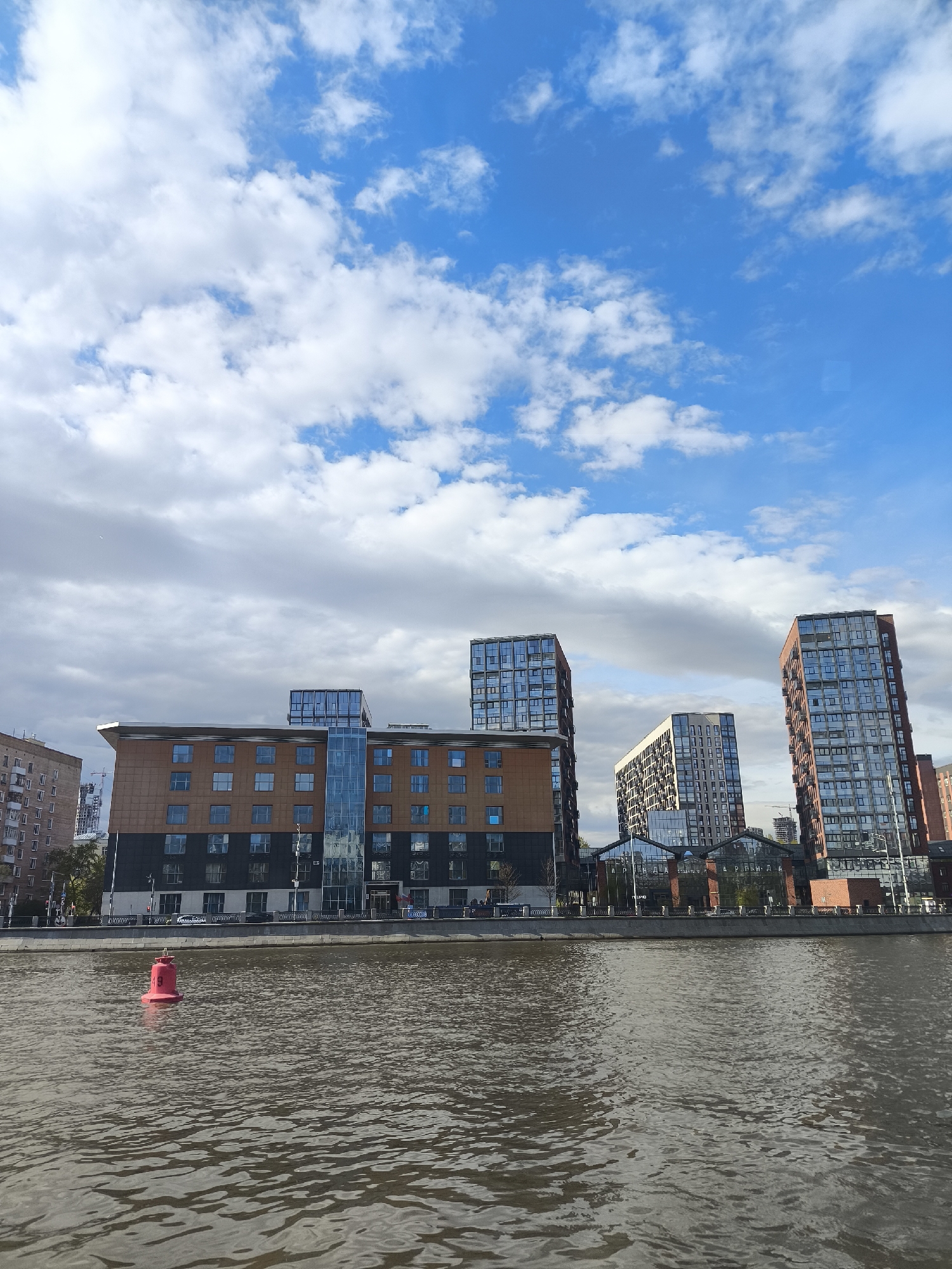 The image depicts a waterfront scene in Moscow, Russia, showcasing a mix of architectural styles. In the foreground, a body of water reflects the daylight, with a red navigational buoy marking a channel. The near shoreline is lined with modern buildings, including a prominent structure with brown and black cladding and a glass-enclosed section. Further behind this building are several high-rise buildings with reflective glass facades, indicative of modern residential or commercial developments. The sky above is partly cloudy with patches of blue, suggesting a fair weather day. No people are clearly visible in the scene.