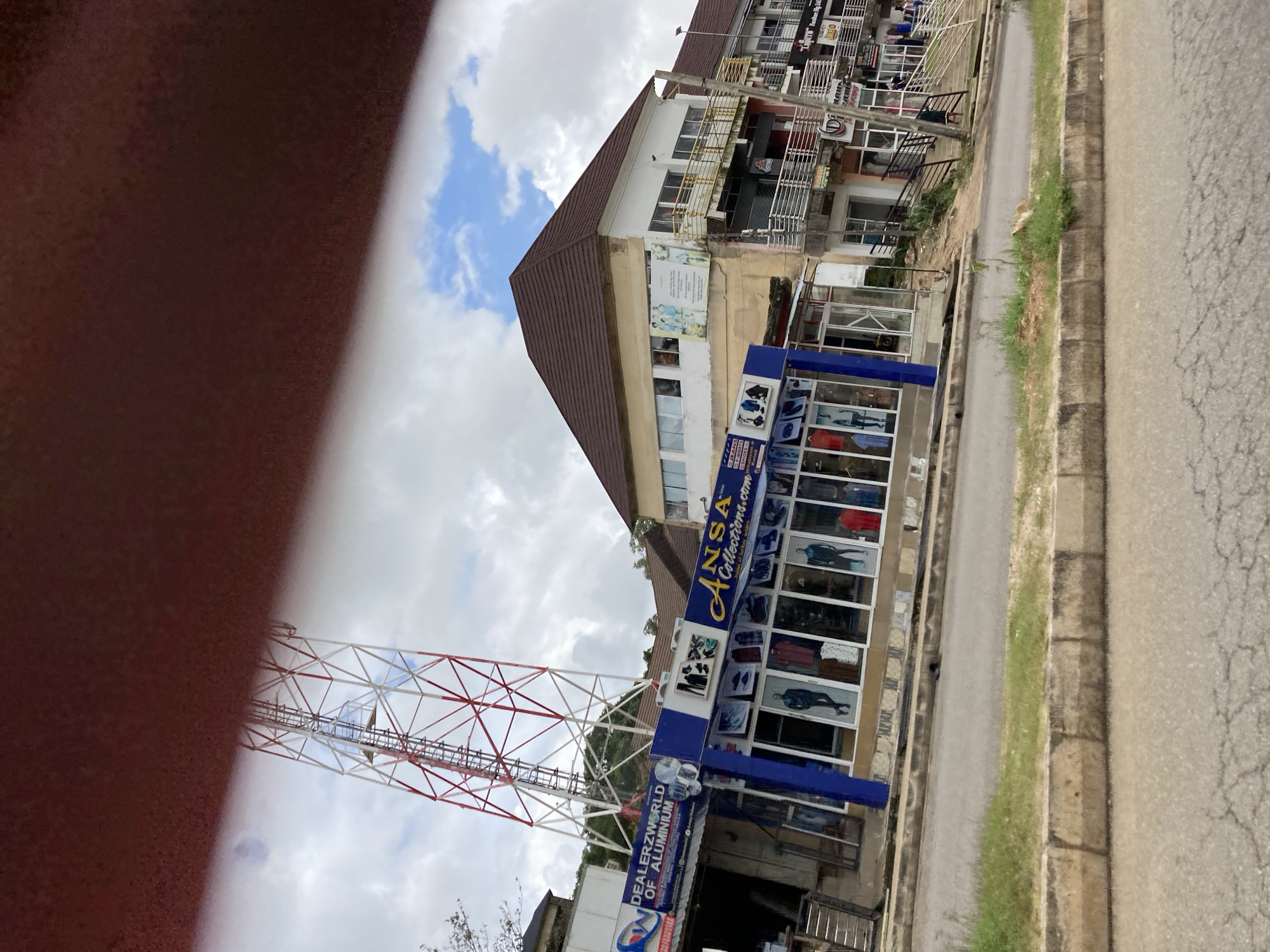 The image captures a street scene in Jos, Nigeria, featuring a row of commercial establishments and a prominent telecommunications tower. The foreground shows a cracked asphalt road bordered by a concrete curb and a strip of patchy green grass. A telecommunications tower, painted with red and white diagonal stripes, dominates the upper left portion of the frame, leaning slightly.
Below the tower, several shops are visible. One shop, identified as "DEALERZWORLD OF ALUMINIUM," displays signage and what appears to be aluminum products. Adjacent to it is "ANSA Collections.com," a clothing store with mannequins dressed in various garments displayed in its large glass windows. Other businesses with signage such as "D'JEANS" and "LIGHTS Furniture Nig Ltd" are also present in the wider storefronts. The buildings are multi-story structures with balconies and multiple windows. The sky is cloudy with patches of blue. Several people are seen walking in front of the shops and on the elevated walkways. The overall scene depicts a busy commercial area within Jos.
