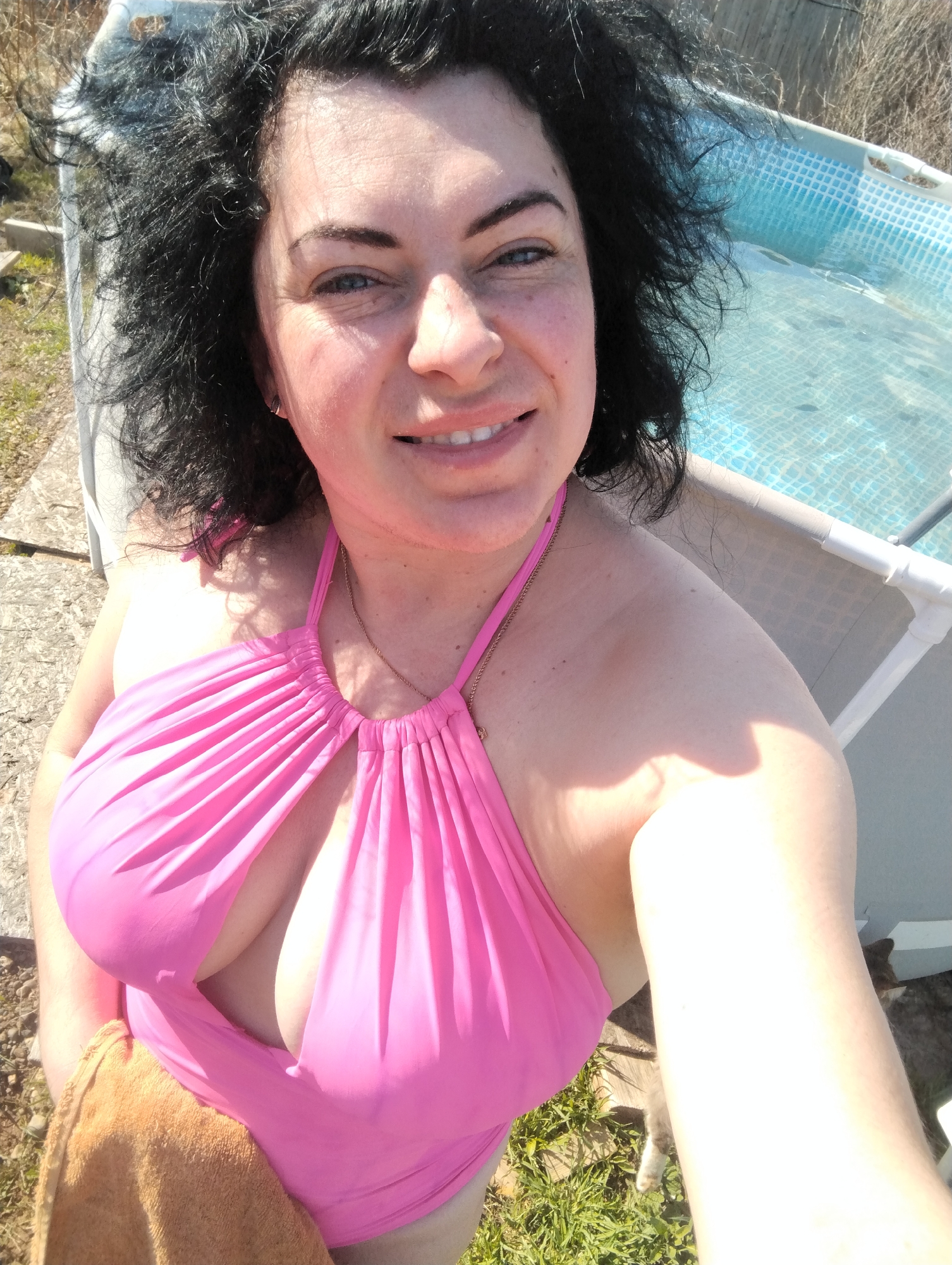 A woman with dark, curly hair and blue eyes is captured in a close-up selfie, smiling directly at the camera. She has fair skin and is wearing a bright pink halter-neck swimsuit with a gathered front. A delicate gold chain is visible around her neck. A brown towel rests on her lap.
The setting is outdoors on a clear, sunny day, indicated by the strong, direct sunlight and visible shadows. Behind her, to the right, is a portion of an above-ground swimming pool with a blue and white patterned liner visible through the clear water. To the left and in the lower background, dry grass and dirt or gravel ground are visible, suggesting a natural or semi-natural outdoor environment. The presence of a light-colored chair frame can be seen behind her shoulder. The scene appears to be taking place in Grishanki, Russia. There is no visible text in the image.
