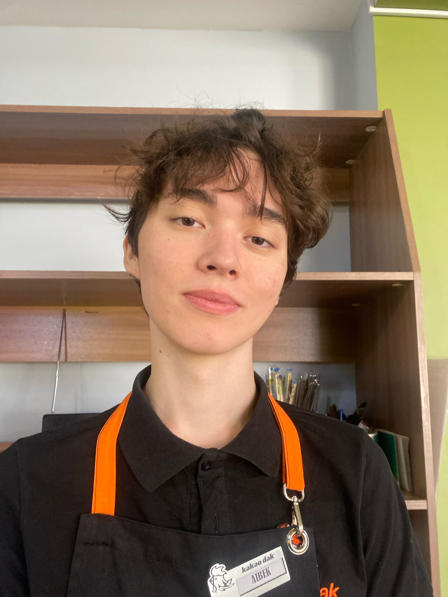A medium shot, eye-level, selfie-style photograph shows a young person with wavy brown hair, wearing a black polo shirt and a black apron with orange straps. The person has light skin, brown eyes, and is looking slightly to their right with a faint smile. A name tag on the apron reads "kakao dak AIBEK". The background consists of wooden shelves. The shelves appear to be in an indoor setting, possibly a cafe or a retail establishment, given the apron and the name tag. The lighting is bright and even. The overall impression is of a candid snapshot taken by an employee at their workplace. The image does not contain any information that would help identify the city of Astana, Kazakhstan.
