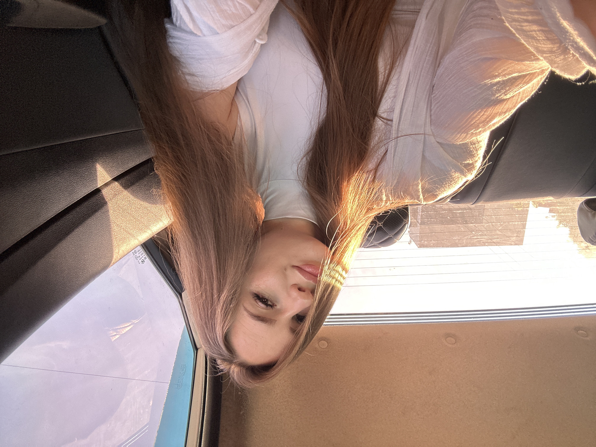 A young woman with long, brown hair is pictured inside a vehicle. She is wearing a white turtleneck top and a sheer white outer layer. The setting sun casts a warm glow on her face and through the rear window of the car. The interior of the vehicle appears to be a backseat, with dark upholstery. The overall mood of the image is soft and illuminated.