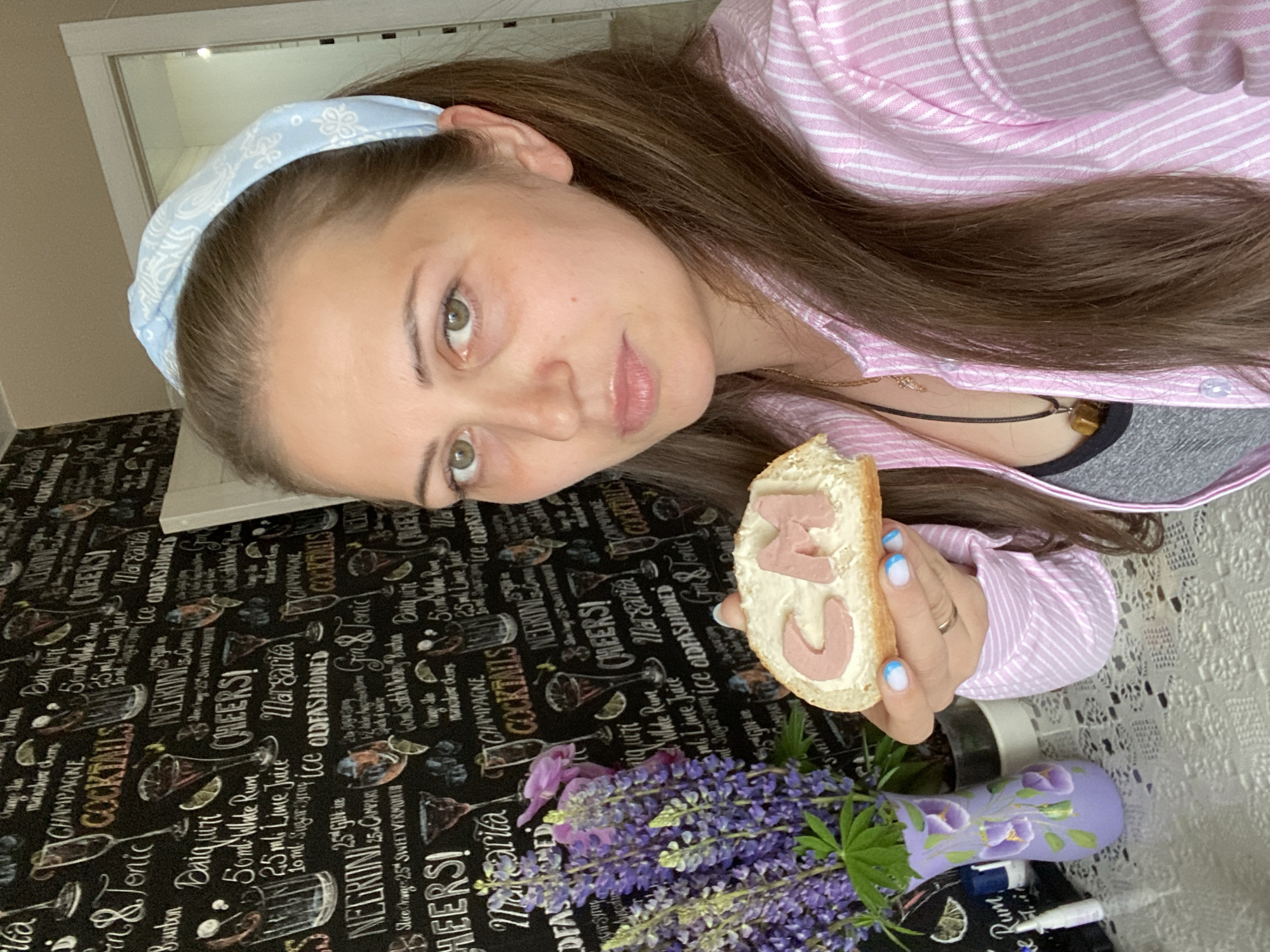 A woman with brown hair and light green eyes, wearing a pink and white striped shirt and a light blue patterned headband, holds a slice of bread with butter and cut-out letters forming "CM". She is positioned in front of a black wall with a pattern of cocktail glasses and names. To her left is a vase with purple lupine flowers. The background suggests an indoor setting, possibly a kitchen or living room. The location is stated to be Марковский, Russia, though this cannot be visually confirmed from the image itself.