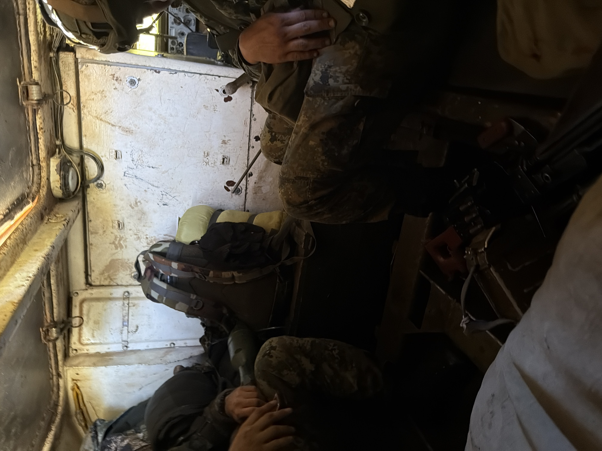 Inside a dimly lit, cramped compartment, likely the interior of a vehicle, two individuals are seated. One person on the left, wearing camouflage pants and a dark jacket, is partially visible, with their hands resting on their lap. They are holding a cylindrical object, possibly a scope or a large flashlight. Next to them is a camouflage backpack. On the right, another person is seated, also in camouflage pants and a dark uniform, wearing a helmet with goggles. Their hands are resting on their leg. The interior walls are panelled and appear worn, with various fixtures and wires visible. There are several bags and equipment within the compartment, including a rifle lying on the floor in the foreground. The overall impression is one of a military or tactical environment. The location is not discernible from the visual information in the image.