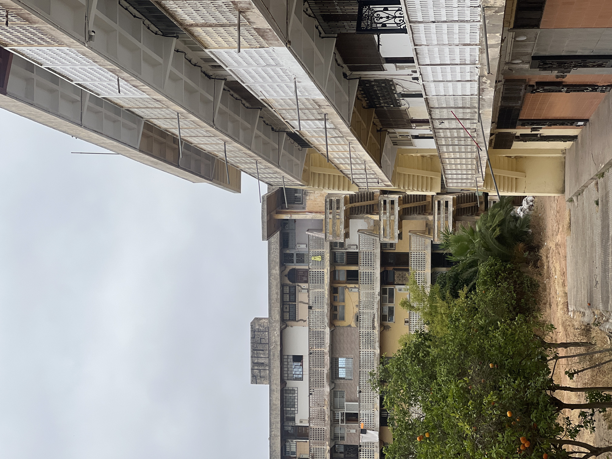 The image captures a dense urban residential area, likely an inner courtyard or alleyway, framed by multi-story buildings under an overcast sky. The perspective looks upward, emphasizing the height and proximity of the structures.
In the center, a large, multi-story apartment building features numerous windows, some with security bars, and multiple balconies or terraces adorned with patterned screens or railings. The building's facade displays a mix of muted yellow, beige, and brown tones, suggesting age and varied construction. Small details like air conditioning units are visible, and a small yellow piece of laundry hangs from one balcony.
Overhead, spanning from the upper right portion of the frame, are several levels of light-colored, grated structures, appearing as extended walkways, sunshades, or part of higher-level access, supported by slender poles. A small security camera is mounted on one of these upper structures. External staircases can be observed between buildings in the background.
At the bottom right, the ground consists of a paved section transitioning to a dry, earthy area. Several trees punctuate the scene, most notably a vibrant green tree laden with visible oranges, along with some palm trees.
The sky is a uniform, light grey, indicating an overcast day without direct sunlight. No people are visible, and no specific activity is depicted, contributing to a quiet, lived-in atmosphere. There is no discernible text within the image. The architectural style and vegetation are consistent with urban residential areas in Casablanca, Morocco.