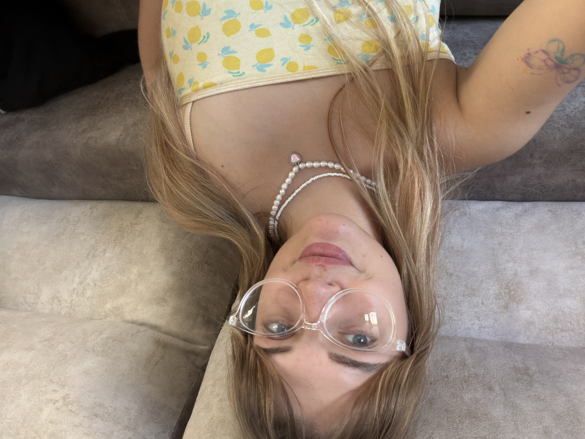A fair-skinned young woman with long blonde hair is lying upside down on a light-colored couch or bed. She is wearing a yellow tank top with a lemon print, clear-framed glasses, and a double-stranded white pearl necklace with a pink heart pendant. A colorful butterfly tattoo is visible on her upper right arm. The photo was taken in an indoor location. There is no information available in the image to confirm it was taken in Большая Шильна, Russia.