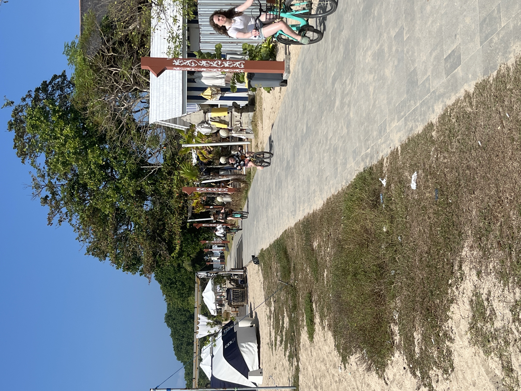In Kuta Selatan, Indonesia, a paved walkway runs alongside a sandy beach, lined with vegetation and a few small, beachfront buildings. The day is sunny with a clear blue sky. Several people are visible. In the foreground, a young woman with curly brown hair rides a teal bicycle with a basket. Further down the walkway, another young person on a bicycle, wearing a floral top and dark shorts, is also riding. In the distance, two adults on bicycles, one wearing a white shirt and the other a darker top, are also pedaling along the path. The beach to the left features white tents and umbrellas, suggesting a commercial or recreational area. The overall scene depicts a leisure activity in a tropical, coastal environment.