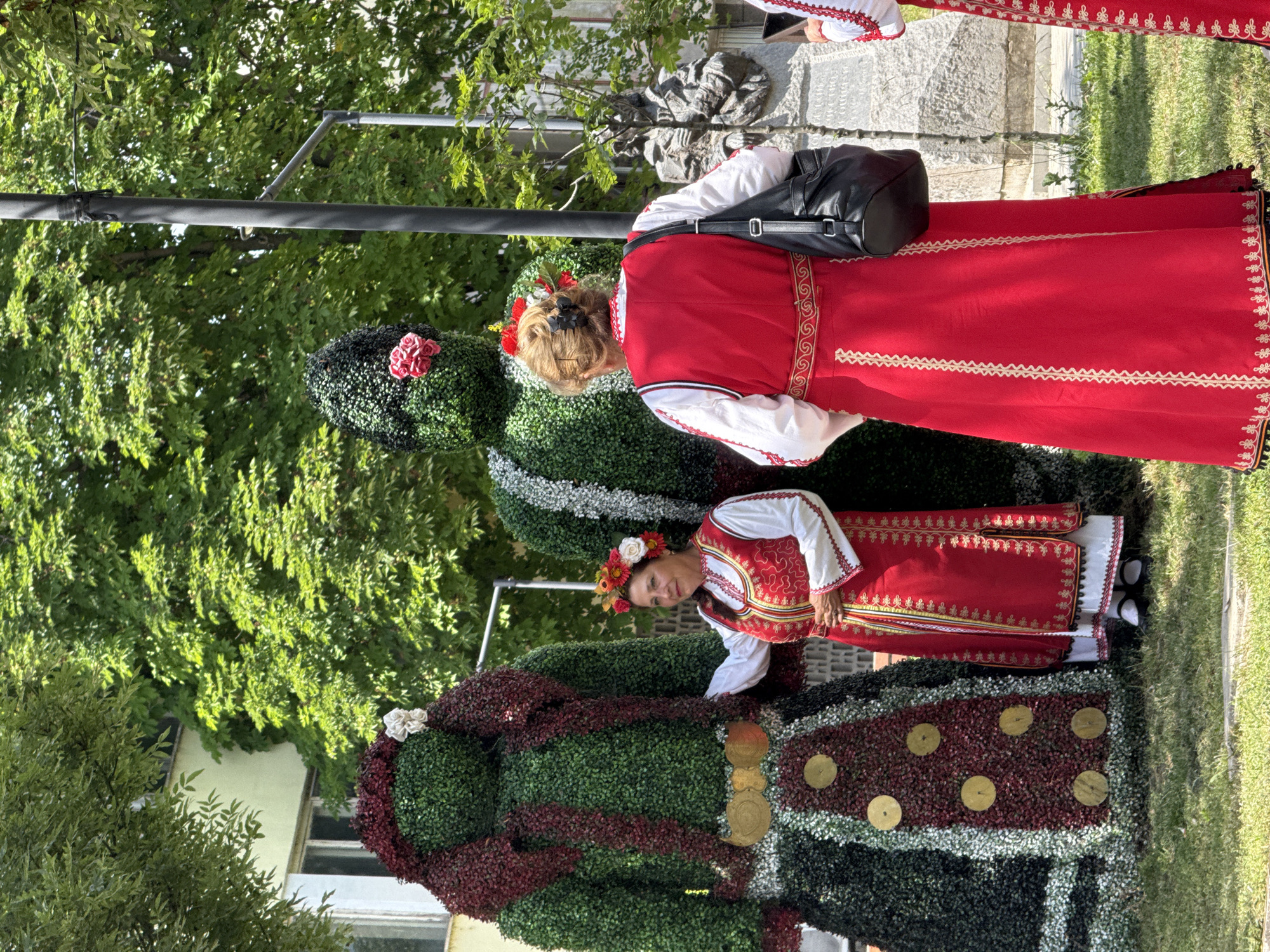 Two women in traditional Bulgarian folk costumes stand in front of two large topiary figures on a sunny day in the city of Pordim, Bulgaria. The topiary figures, which appear to be dressed in clothing made of greenery and burgundy leaves, resemble people. The woman on the left, closer to the camera, is facing the viewer, her right hand resting on the left topiary. She is wearing a white blouse, a red vest embroidered with gold thread, and a red skirt. She has a flower crown on her head. The woman on the right is seen from behind, wearing a red dress with white embroidery and a black purse slung over her shoulder. Her hair is styled with flowers. The figures and women are on a grassy area, with trees and a building in the background. A street lamp and a stone monument are also visible.