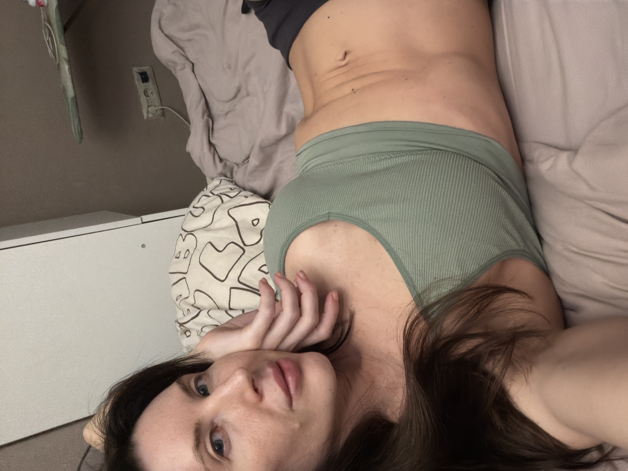 A young adult female is lying on a bed, angled toward the camera. She has dark brown hair, fair skin, and blue eyes. She is wearing a green ribbed tank top and appears to be wearing gray shorts. Her right hand is near her face. The bed is covered in a beige duvet, and a pillow with a black and white geometric pattern is under her head. A white bedside table is visible on the left side of the frame. In the background, a wall with an electrical outlet can be seen. The location is a bedroom, potentially in Perm, Russia, based on the request.