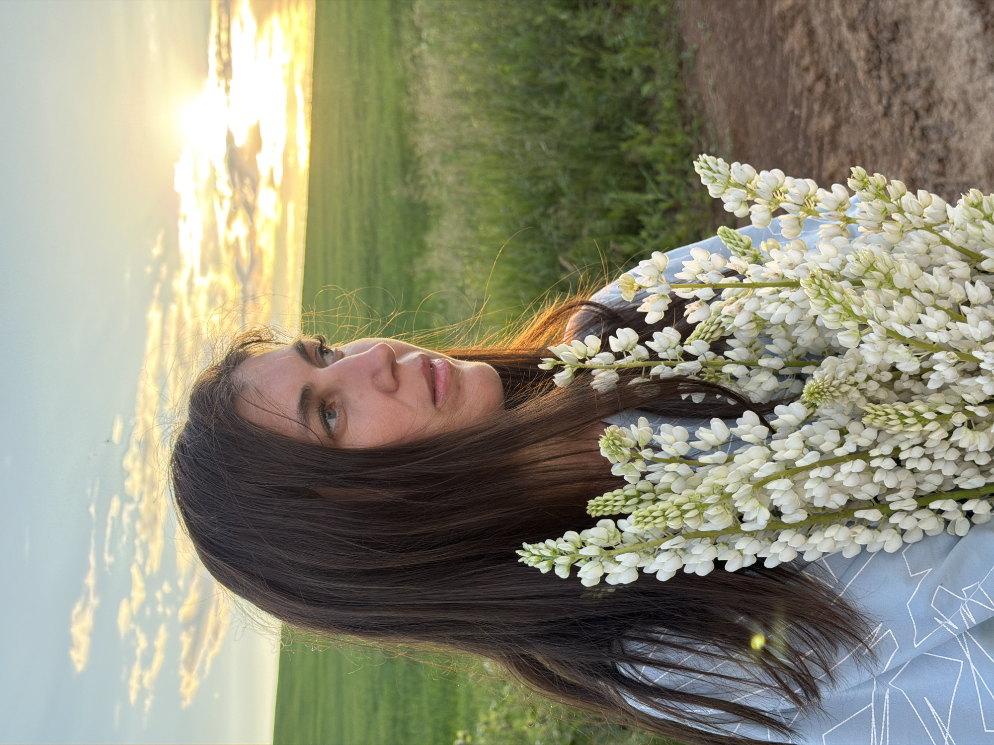 A woman with long, dark brown hair stands in a field at sunset, holding a bouquet of white lupine flowers. She is wearing a light blue shirt with a geometric pattern. The background consists of a green field and a sunset sky with clouds. The location is Vassyata, Russia.