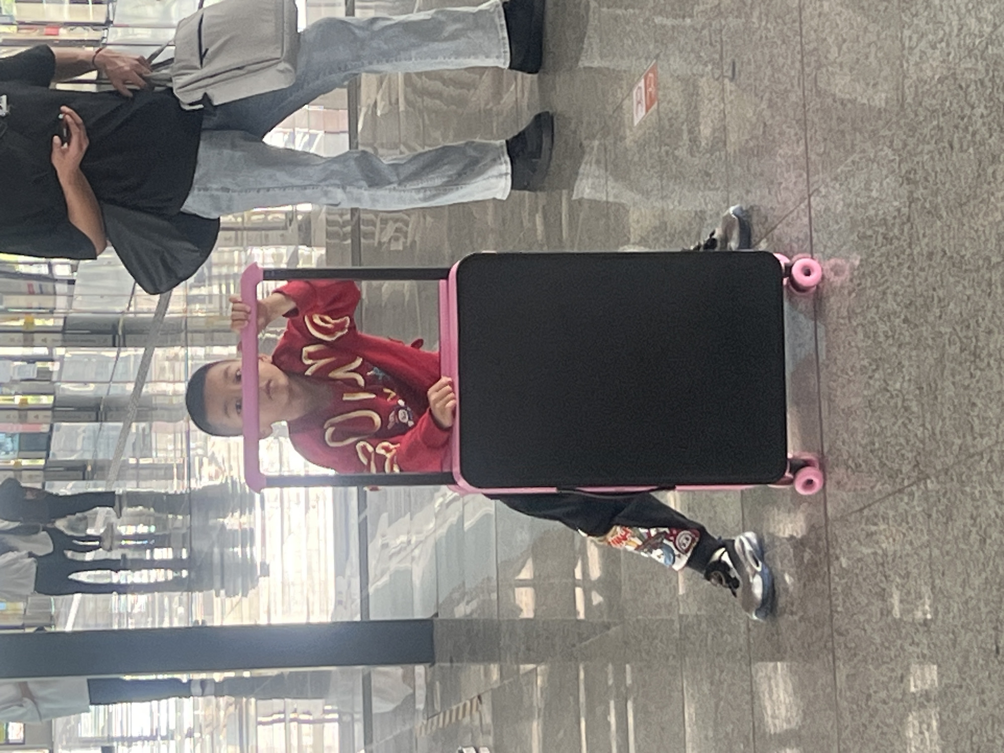 This portrait-oriented photograph captures a child and an adult in an indoor public space, likely a modern facility such as an airport, train station, or shopping mall in Jiangjin, China. The setting features a light-colored tiled floor and tall, reflective wall panels or pillars that create distorted reflections of the environment and unseen individuals.
In the foreground, a young boy, appearing to be between five and eight years old, is seated inside a pink and black wheeled device that resembles a child's luggage or a specialized stroller. He wears a red long-sleeved sweatshirt adorned with white graphics and dark trousers with colorful, cartoon-like patches on the lower legs. His face is visible above the top edge of the carrier, and he looks towards the viewer with a calm expression, gripping the pink handle. He is also wearing light-colored sneakers with dark accents.
Standing slightly behind and to the left of the child is an adult, whose upper body and face are not fully visible. The adult is dressed in a black t-shirt, light blue jeans, and dark shoes, and carries a light gray shoulder bag. They appear to be accompanying or supervising the child.
On a reflective surface in the background, to the right of the adult's legs, there is a small red rectangular sign with indistinct white characters, which are too blurry to decipher. The lighting is bright and even, typical of a well-lit indoor environment. The activity suggests travel or leisure within a public venue.
