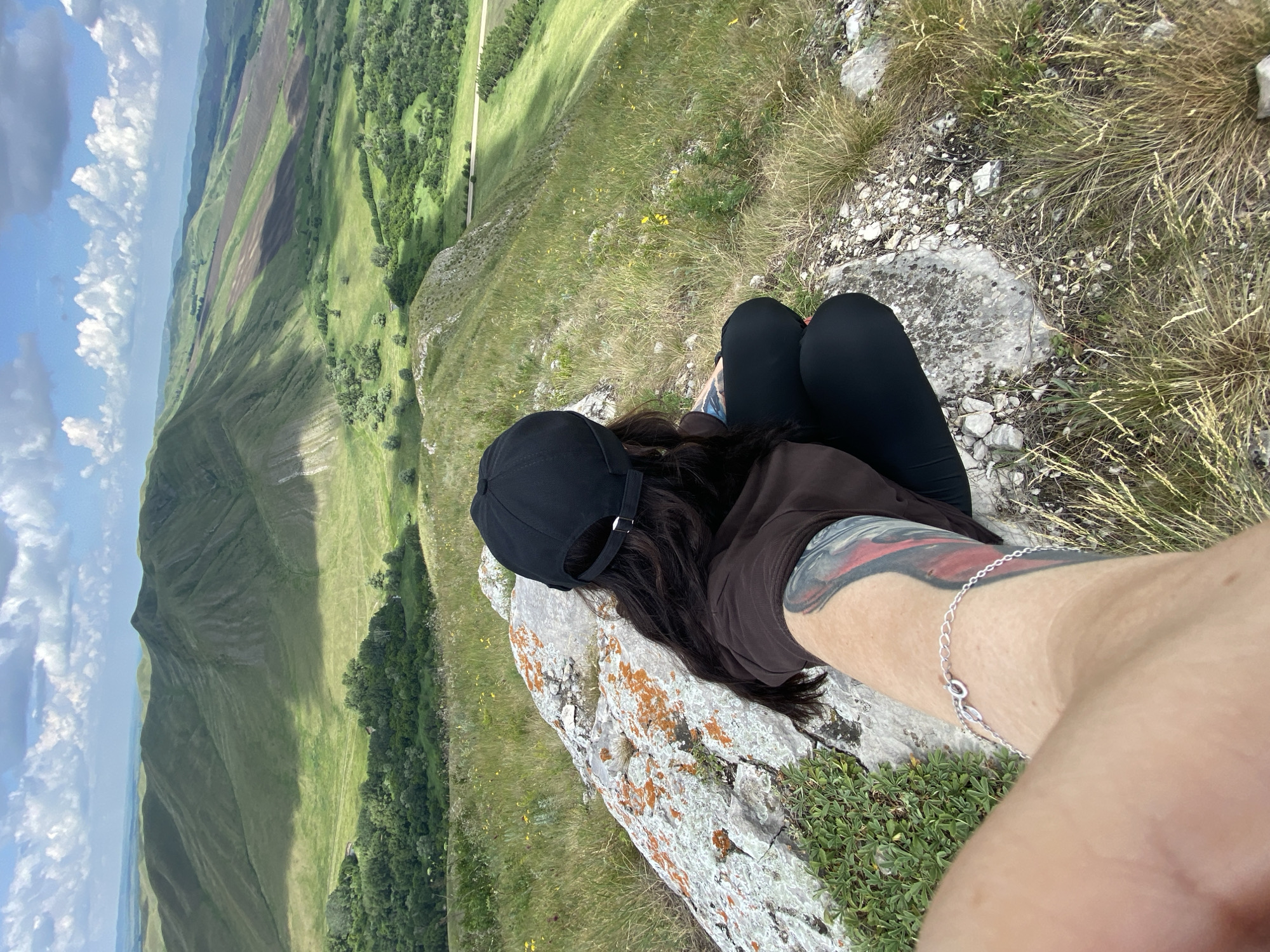 The image captures a scenic view of rolling green hills and valleys under a partly cloudy sky. In the foreground, a person wearing a black baseball cap and a brown hoodie is seated on a large rock, facing away from the camera. The person is looking out at the expansive landscape. The individual's left arm is visible, displaying a tattoo and a silver bracelet. The rock has patches of orange lichen on its surface. The surrounding ground is covered with dry grass and smaller rocks. The background reveals a vast agricultural landscape with fields and scattered trees, interspersed with the undulating terrain of the hills. The location is identified as Petrovskoe, Russia.