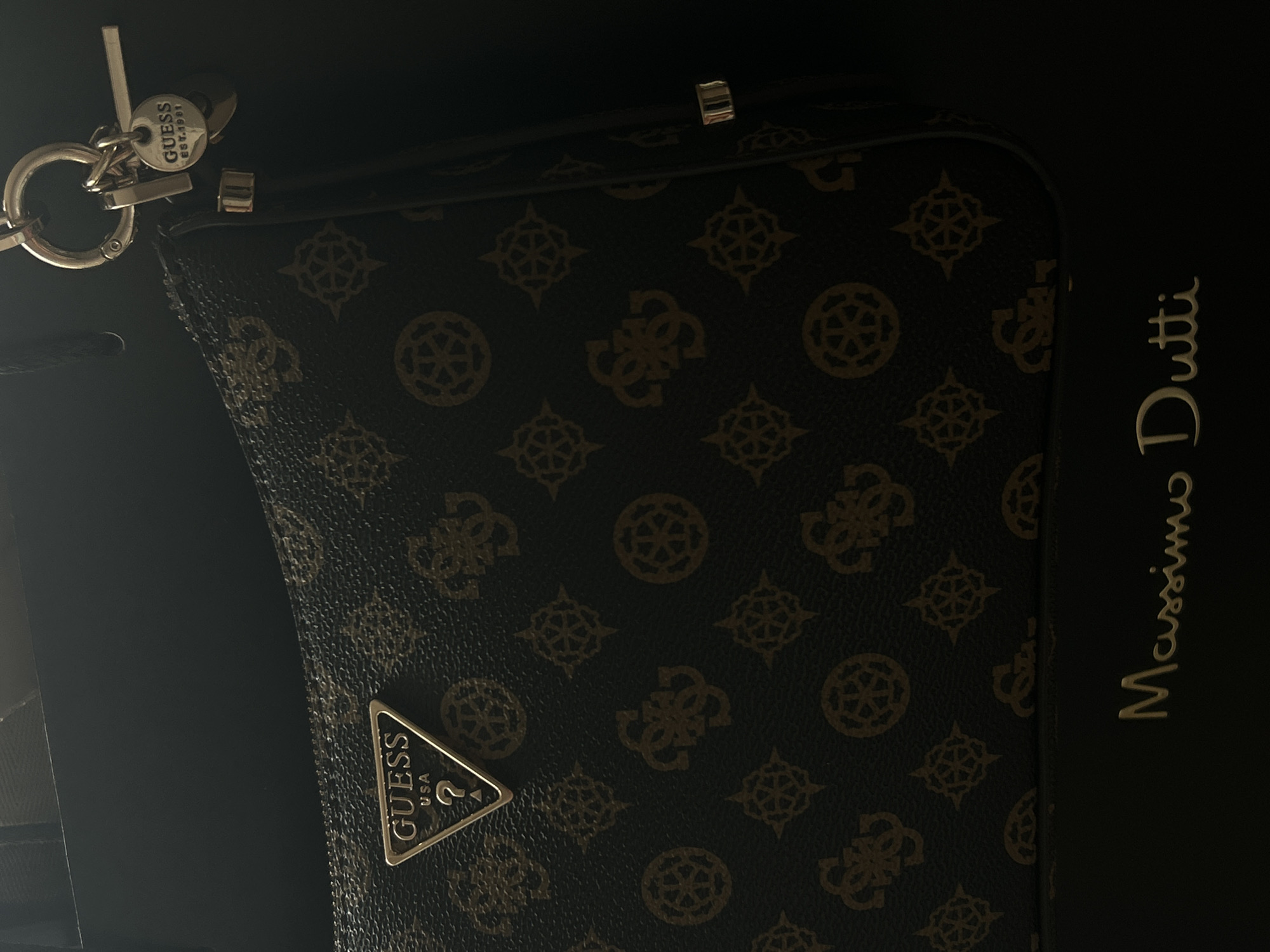 The image depicts a Guess handbag with a brown monogram pattern, placed next to a black Massimo Dutti box. The handbag has a metal logo on its front and a keychain attachment at the top. The lighting is dim. The location is not apparent.