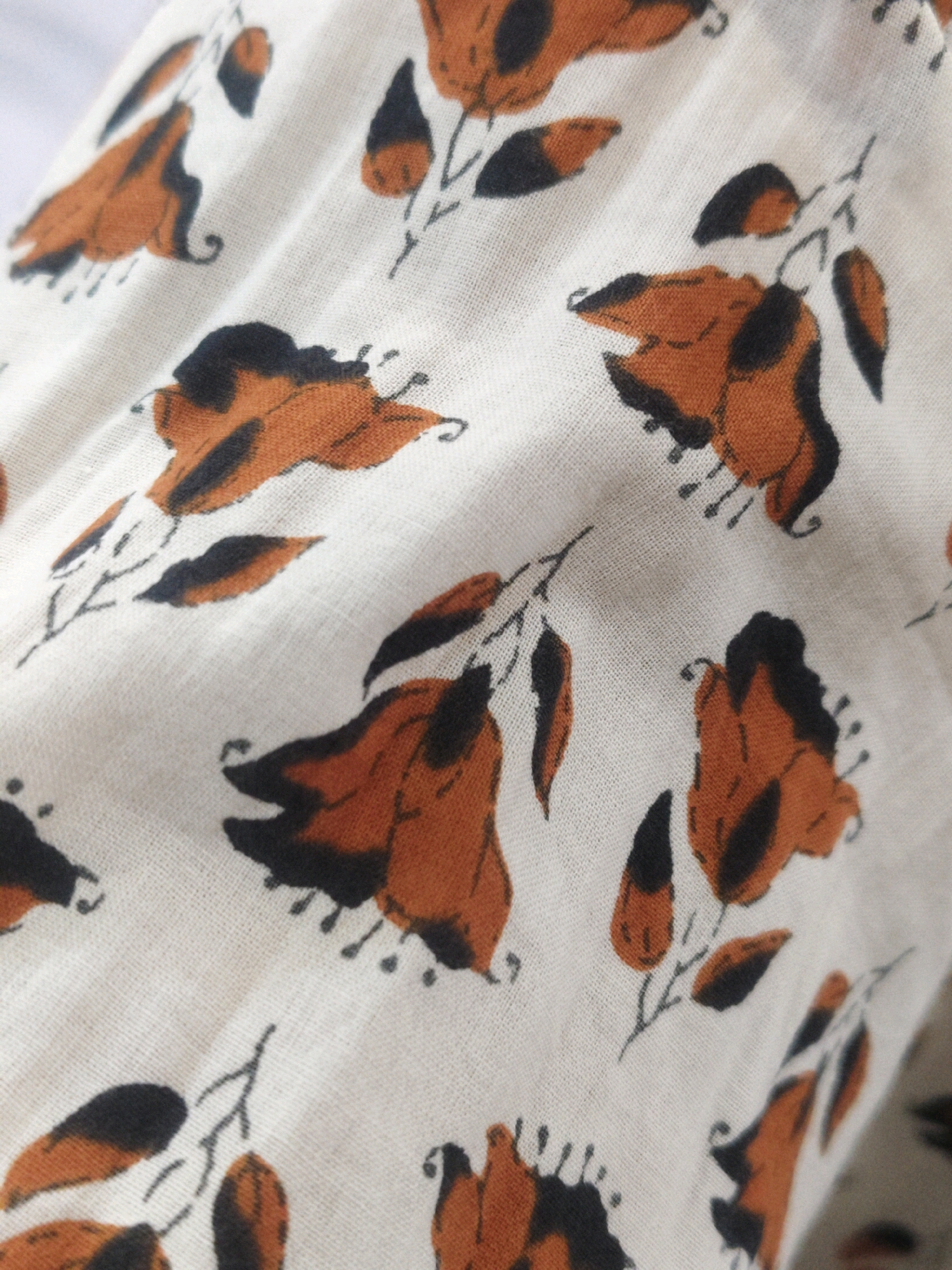 The image is a close-up, detailed shot of a fabric with a floral pattern. The fabric appears to be a light color, possibly off-white or cream, with a visible texture of threads. The pattern consists of stylized flowers and leaves rendered in shades of burnt orange and black. The flowers are abstract, with irregular petal shapes, and the leaves are elongated and pointed. The brushstrokes are visible, giving the pattern a hand-painted or block-printed feel. The fabric is slightly creased and folded, indicating it is likely a piece of clothing, drapery, or material. There are no people, animals, or specific landmarks visible in the image. The context provided about the location in Sumerpur, India, is not visually supported by the content of the image itself.