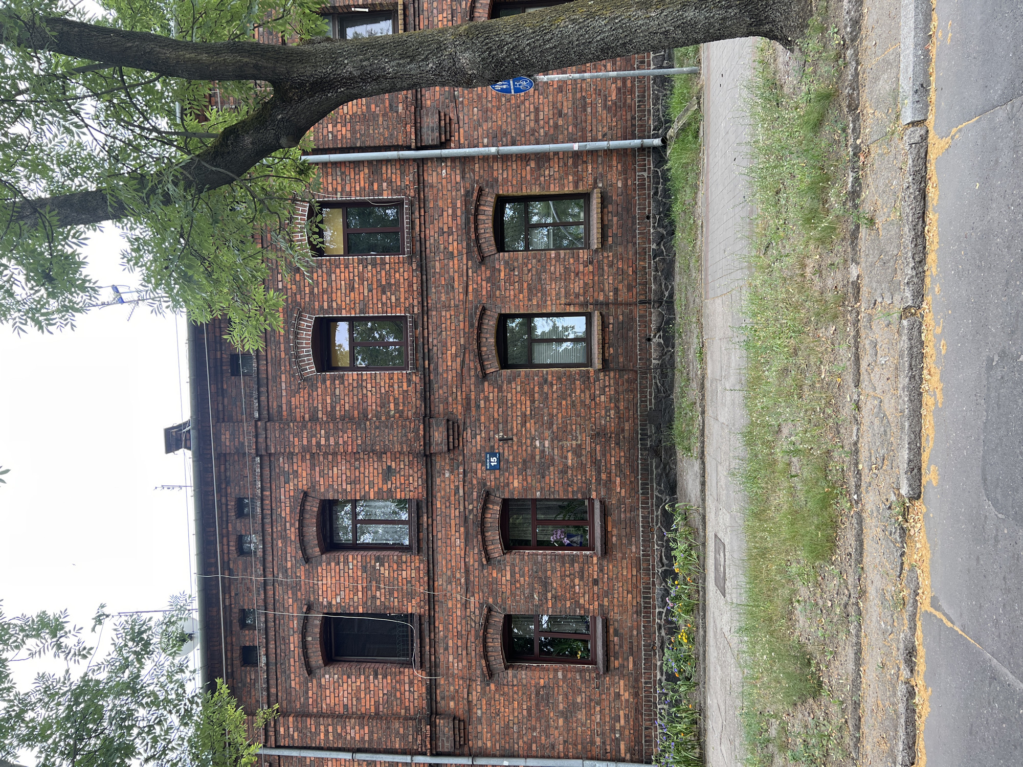 This is a medium-shot, eye-level, exterior, daytime photograph of the brick facade of a multi-story building. The building has at least two floors visible, with several windows on each. The windows are rectangular with dark frames. The building appears to be made of red brick. In front of the building, there is a sidewalk and a patch of grass. A tree with green leaves is in the foreground on the right side of the image. The sky is overcast. There is a street sign that says "Konopnicka 15" on the wall of the building. This is the building at Konopnicka 15 in Żyrardów, Poland.