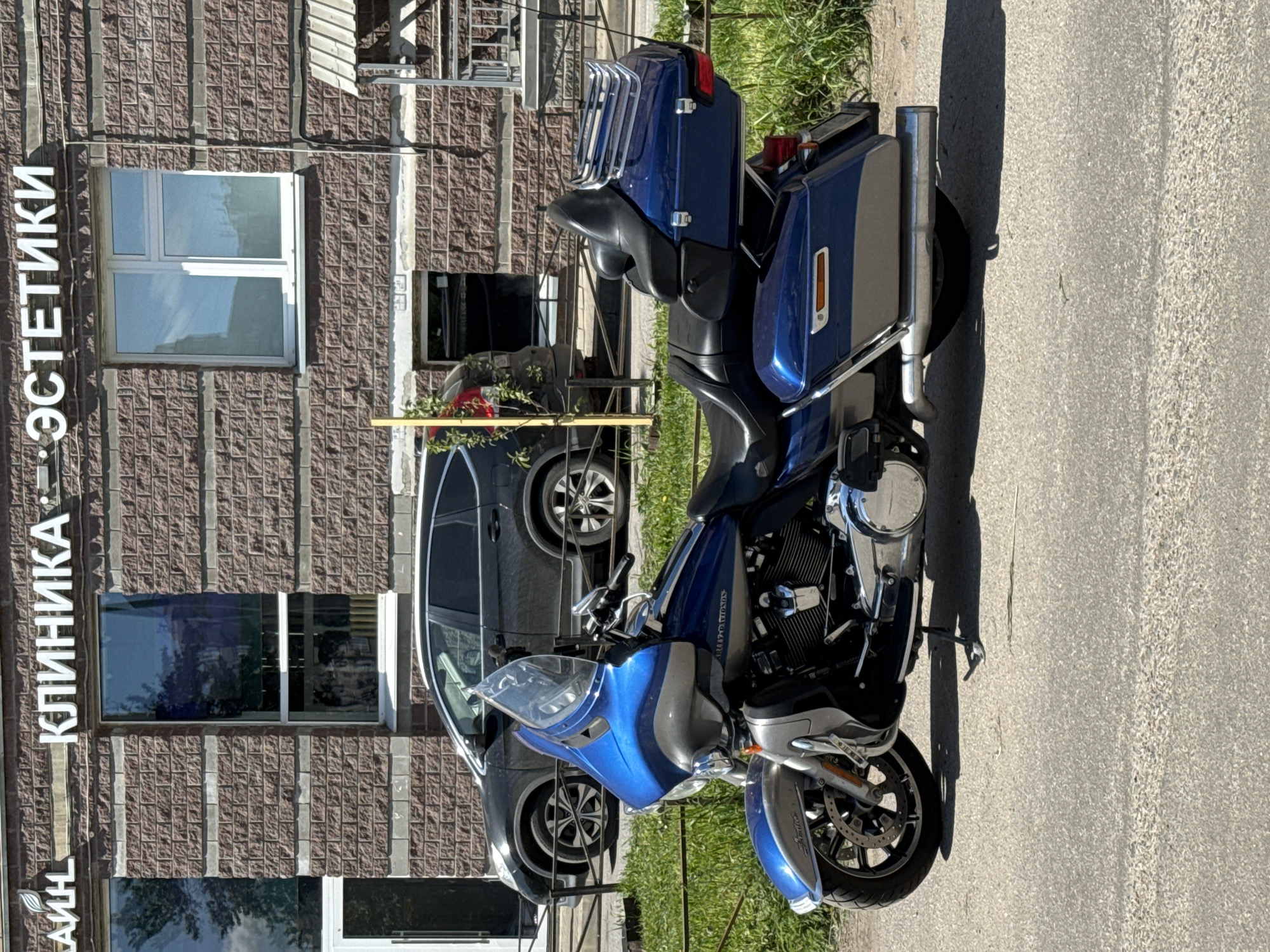 A bright, sunny outdoor scene captures a blue and silver touring motorcycle, likely a Harley-Davidson, parked on a paved surface. The motorcycle is positioned on the right side of the frame, facing left, with its detailed engine, chrome accents, large saddlebags, and rear luggage rack clearly visible. Long shadows cast by the motorcycle suggest either morning or late afternoon sunlight.
In the background, a multi-story building with a reddish-brown brick-patterned facade stands, featuring several windows reflecting the sky and surroundings. Prominently displayed vertically on the building's left side is white text in Cyrillic, reading "КЛИНИКА ЭСТЕТИКИ," which translates to "Aesthetics Clinic." Behind the motorcycle and partially obscured by a railing and some foliage, a dark-colored SUV or car is also parked. A strip of green grass separates the paved area from the building. The image presents a clear day in what appears to be an urban or suburban setting in Sinyavino, Russia.