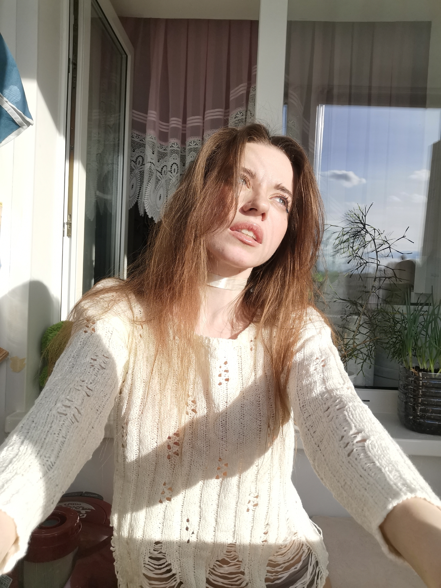 A woman is the central subject, wearing a white sweater with distressed details and a light-colored choker. She's looking towards her right, with light entering the scene from her left, casting shadows on her sweater. Behind her is a window with visible curtains and exterior scenery, including greenery and a view of the sky. To the right of the woman, there is a plant in a dark-colored pot on the windowsill. In the lower left of the frame, there is part of what appears to be household equipment or furniture, partially obscured. The location is Unknown, Unknown.