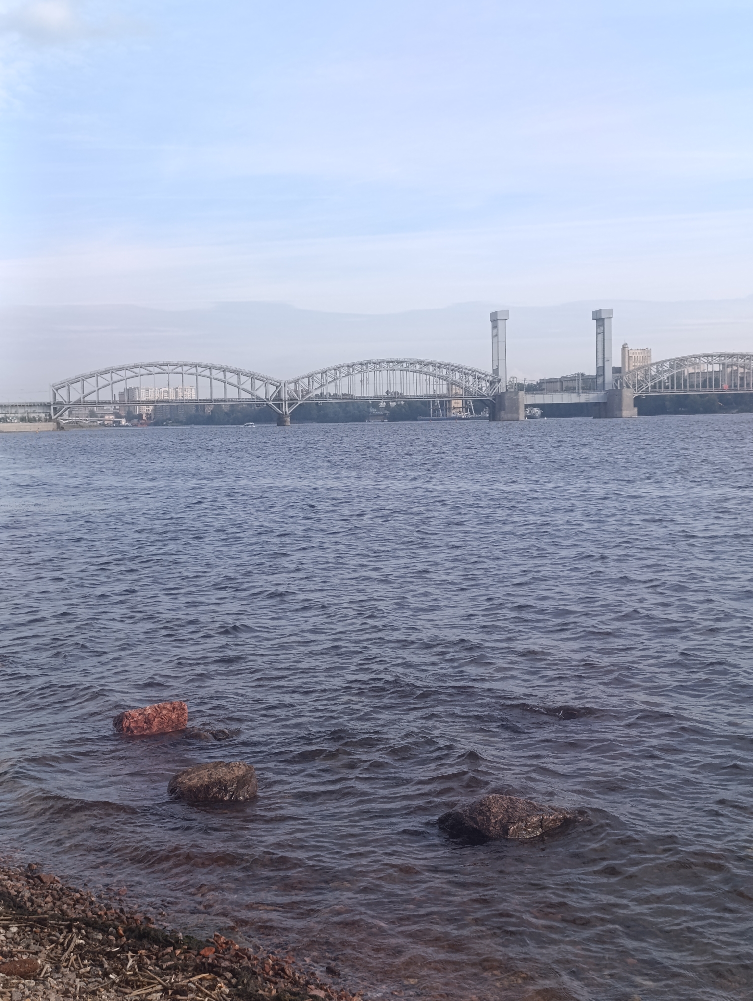 The image captures a wide shot of the Neva River in Saint Petersburg, Russia, on a moderately overcast day. In the foreground, a rocky shoreline meets the dark, rippling water, with three stones partially submerged. Across the river, a complex network of bridges dominates the horizon. The most prominent structure is a large, arched railway bridge, possibly the Bolsheokhtinsky Bridge, with its distinctive twin towers, known as the "gate towers." To the left and right of this central structure, additional bridge spans and industrial buildings are visible, suggesting a significant transportation hub. A few small boats can be spotted on the water, and the sky is a pale, hazy blue with wispy clouds. The scene conveys a sense of urban industrial landscape combined with the vastness of the river.