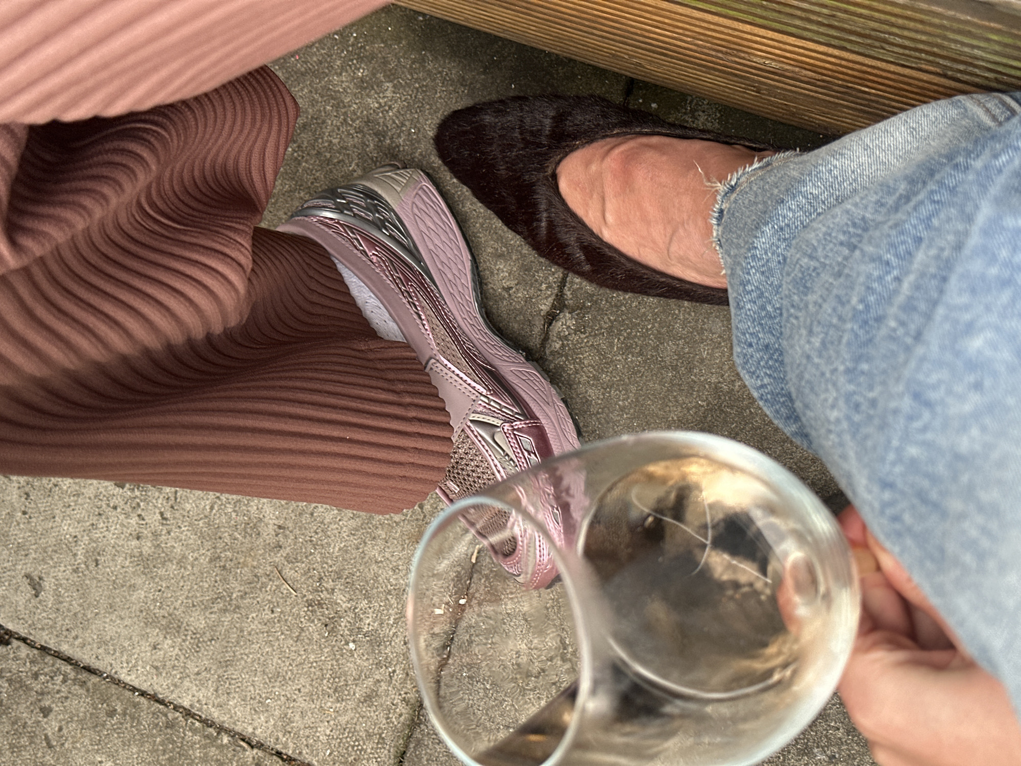 The image shows an overhead view of three people's feet and legs. One person is wearing a brown, pleated dress and pink sneakers. Another person is wearing blue jeans and brown, furry flats. A third person, holding a wine glass, is partially visible in the lower right corner, also wearing jeans. The setting appears to be outdoors, on a concrete surface next to a wooden structure, possibly a bench or fence. The exact location within the Folkestone and Hythe District, United Kingdom, is not discernable from the image.