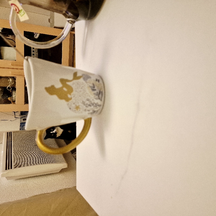 A tilted, close-up shot shows a white mug with gold accents and a mermaid design. The mug sits on a white surface. A glass teapot is seen behind it. An air conditioner and wooden furniture are in the background to the left. The lower part of the image shows a section of tan carpeting or furniture.