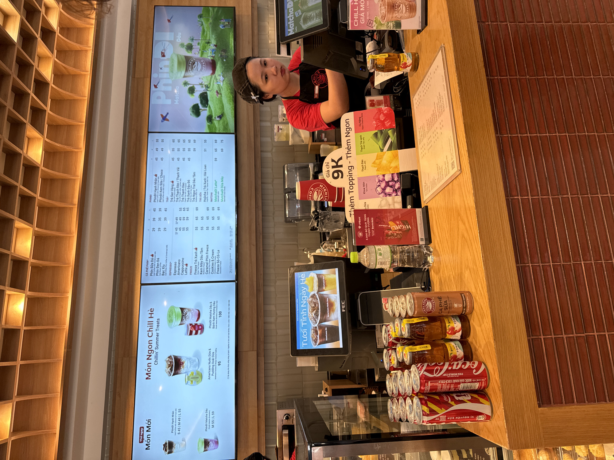 The image shows a woman working at a counter in a coffee shop. She is wearing a red shirt and a dark bandana. She is looking at the camera with a neutral expression. The counter is filled with various items, including a computer monitor, cups, bottles of drinks, and cans of soda. There are also menus displayed on the walls and on the counter, showing prices and names of drinks in Vietnamese. The coffee shop appears to be brightly lit and busy. The overall impression is that of a typical day at a coffee shop in Vietnam, likely during the day given the lighting. The drinks menu suggests a focus on coffee and tea beverages, with some summer treats advertised. The presence of Coca-Cola cans and a "FIFA World Cup" advertisement indicates a possible tie-in with the event.
