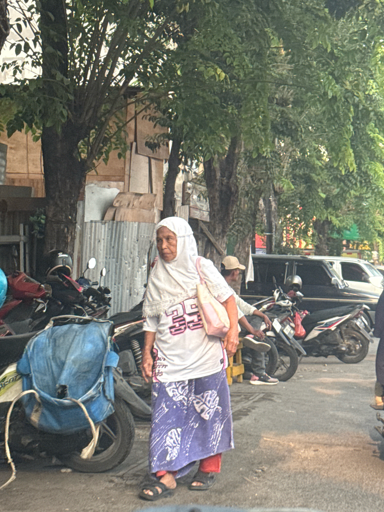 An elderly Indonesian woman wearing a white hijab and a white t-shirt with pink lettering stands on a street in Surabaya, Indonesia. Her t-shirt has the numbers "29" prominently displayed. She is also wearing a long, purple and white patterned skirt and black sandals. She carries a light pink handbag over her shoulder. Behind her, several motorcycles are parked, one of which is covered with a blue tarp. To the right, a dark-colored car is visible, and further back, a man wearing a tan cap and dark pants is seated on a motorcycle. The street appears to be lined with trees and buildings with corrugated metal siding and wooden structures. The overall scene depicts a street view with common elements of urban Indonesian life.