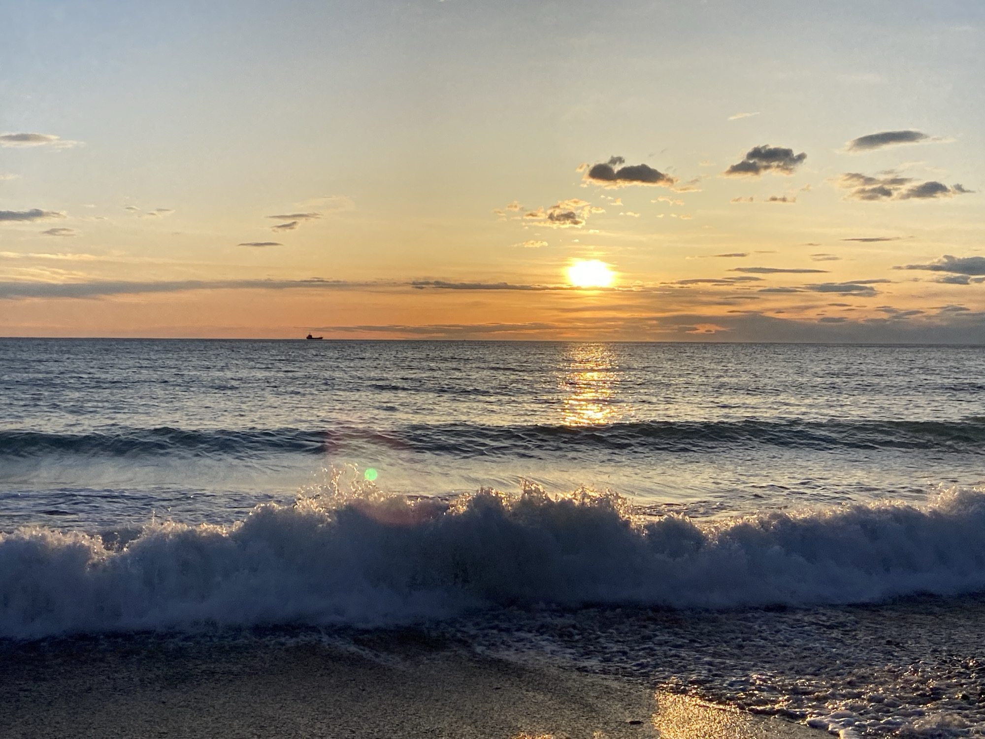 The image depicts a sunset over the Adriatic Sea, likely taken near Bar, Montenegro. The sun is partially obscured by clouds, casting a golden light across the water. A wave is breaking in the foreground, creating white foam. A small vessel can be seen in the distance. The beach appears sandy, with the wet sand reflecting the sunset's glow.