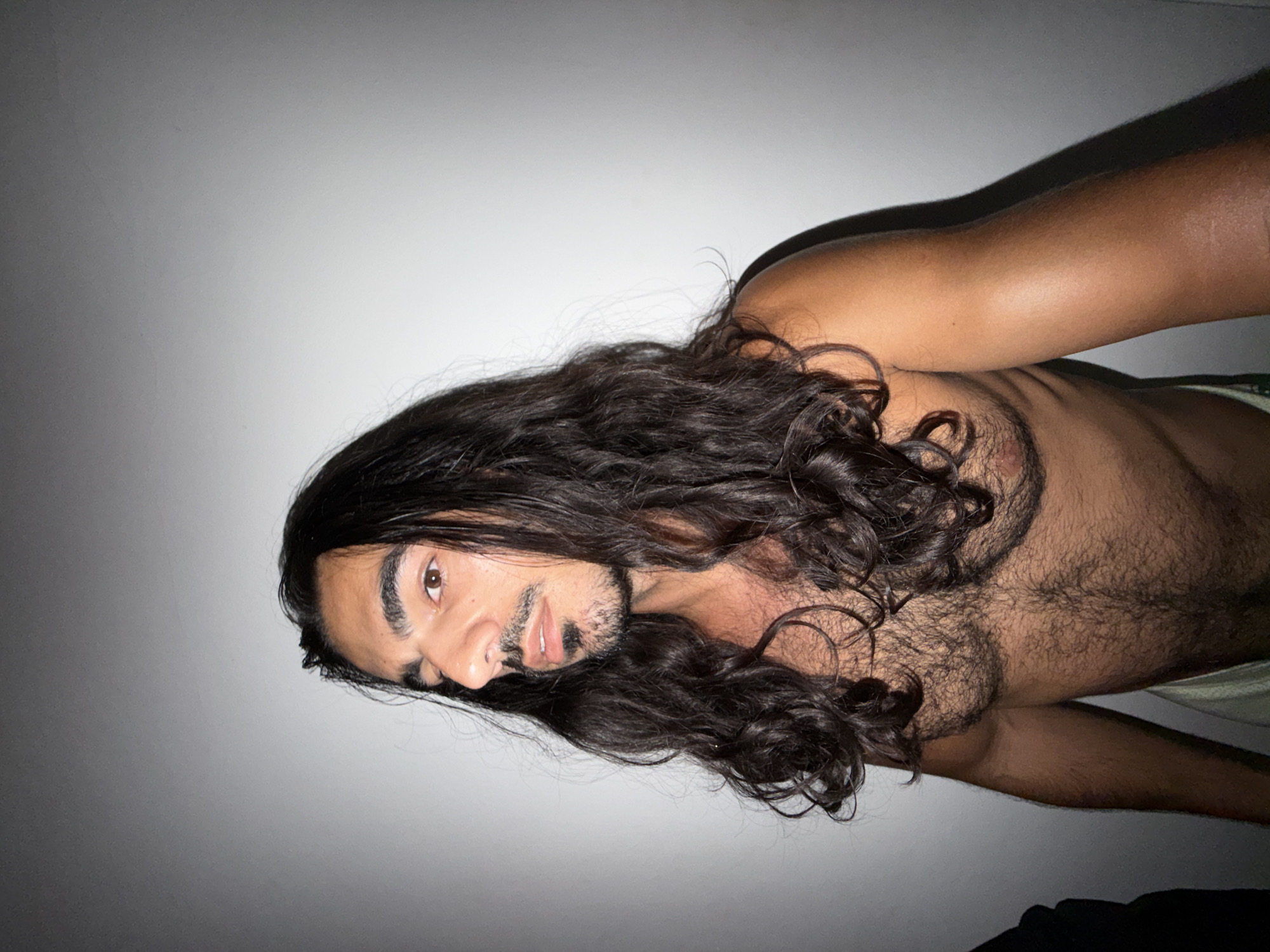 A topless man with long, dark, curly hair and a mustache and goatee is lying down in what appears to be an indoor setting. The background is a plain, light-colored wall. The man is facing the camera and appears to be resting. The location is not discernible from the image. North Kuta, Indonesia, cannot be inferred from this image.