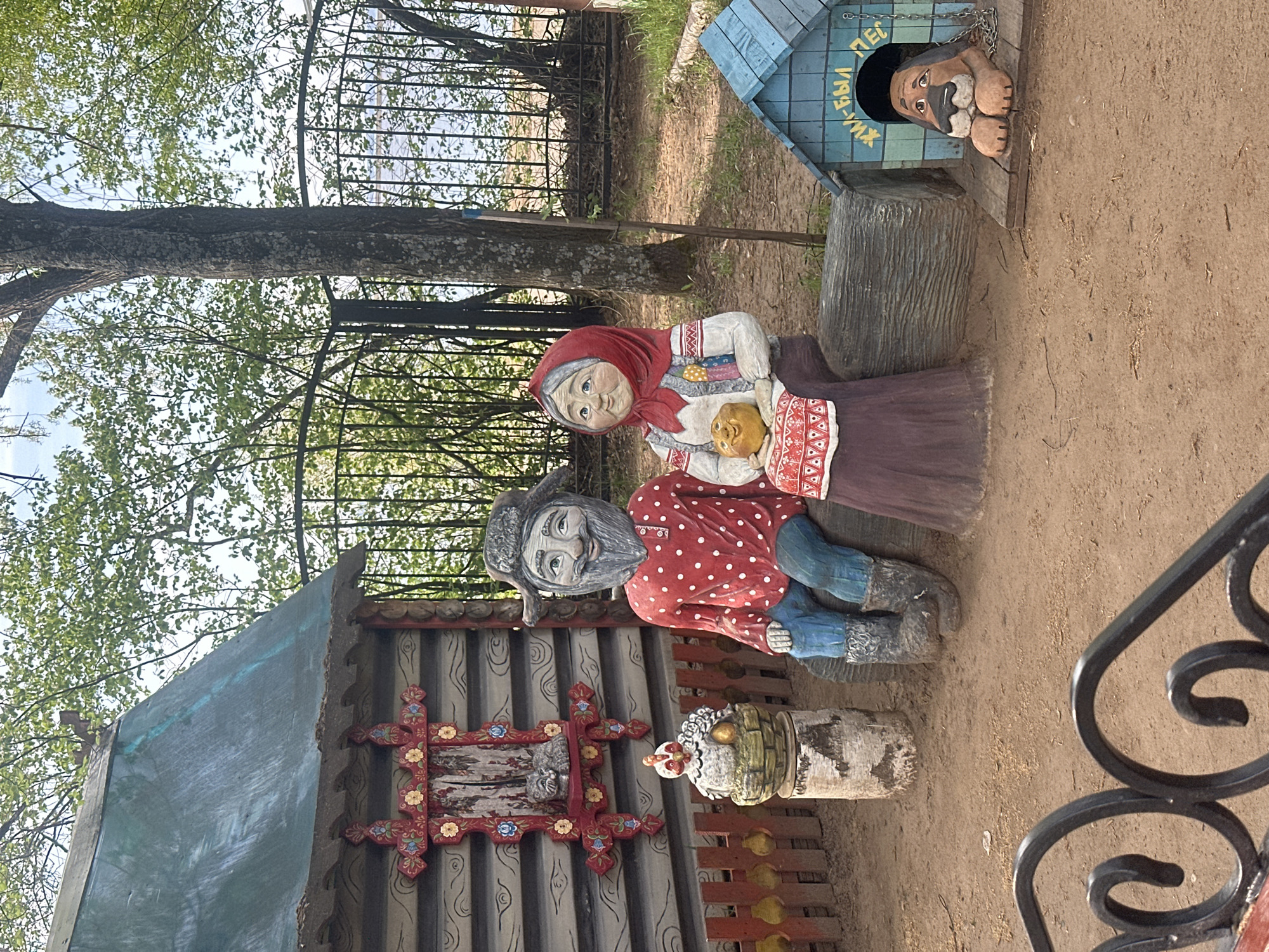 The image shows a collection of wooden folk art sculptures in what appears to be a park or outdoor recreation area. There are sculptures of a man and woman seated together, a doghouse with a dog sculpture emerging from it, and a small structure with decorative elements.
The scene is set on a dirt-covered area, suggesting an outdoor setting. The background features trees and a metal fence, further supporting the park or garden location. The wooden sculptures are painted in bright colors and depict traditional figures and objects.
The location is Chaikovsky, Russia.