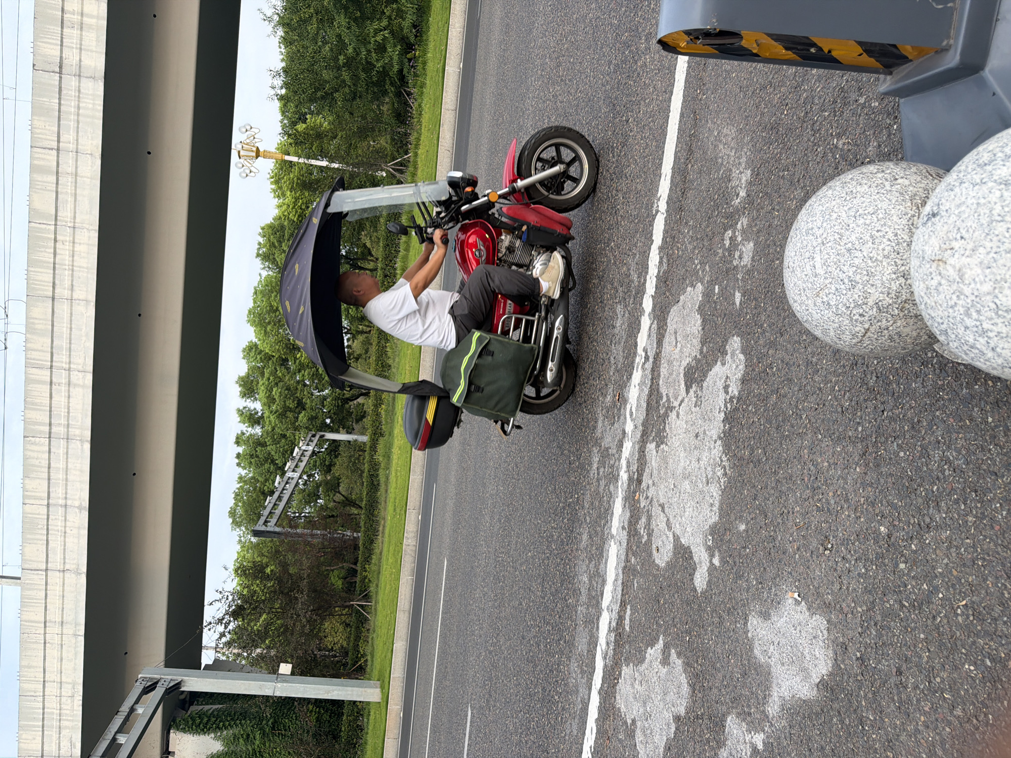 In Jiangjin, China, a man is riding a red motorcycle on a paved road. The motorcycle has a black canopy with a yellow pattern to shield the rider from the sun. The man is wearing a white t-shirt and dark pants. The motorcycle is equipped with a black rear storage box and a green saddlebag. The road is marked with a white line. In the background, there is lush green foliage and trees, with a concrete overpass structure visible above. The scene appears to be a typical street in an urban or suburban area.