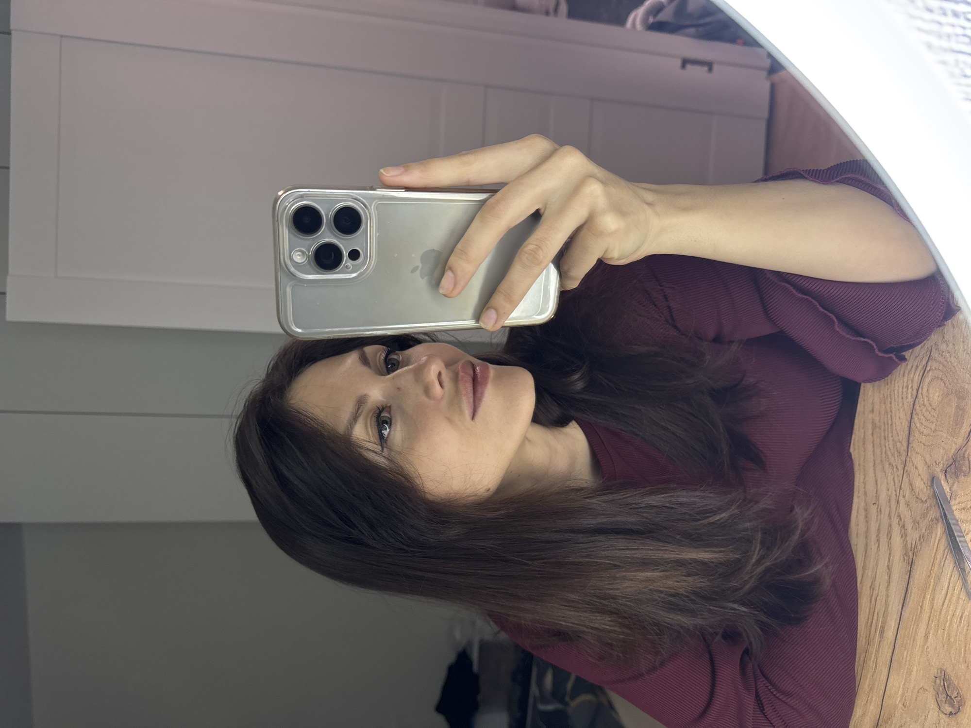 This image is a mirror selfie taken by a woman in Murino, Russia. She is holding an iPhone in her right hand, which features a clear case and prominent triple camera lenses. The phone's "iPhone" logo is visible on its back.
The woman has long, dark brown hair and is wearing a dark red or maroon ribbed top with ruffled cuffs. Her head is tilted slightly upwards, and she has a calm or contemplative expression.
The setting is indoors, likely a room within a home. The background reflected in the mirror includes a light-colored, possibly white, cabinet or wall with horizontal paneling on the left side. To the right, a bright, circular ring light is partially visible, indicating the source of illumination. Below the ring light, a wooden surface or table is present, upon which a pair of tweezers can be seen. The lighting is bright, creating a well-lit environment for the selfie.