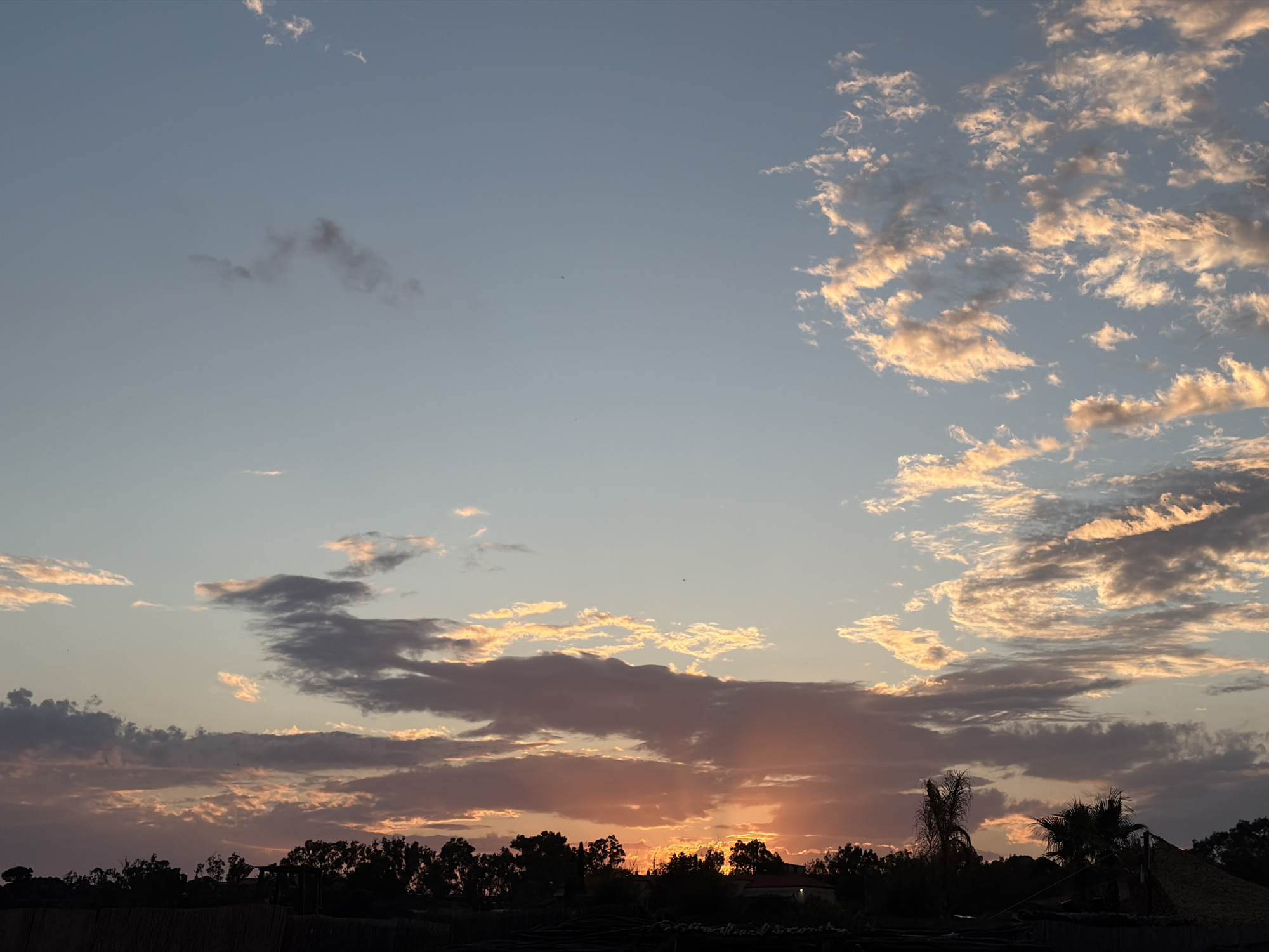 This media file captures a serene sunset over Voula, Greece. The sky dominates the upper two-thirds of the image, displaying a gradient from light blue at the top to warm hues of orange, pink, and purple near the horizon. Scattered clouds are a prominent feature; bright, golden-white cumulus clouds are illuminated by the setting sun on the right side, while darker, elongated clouds stretch across the center and left, some with glowing edges.
The sun is partially obscured by these clouds near the horizon, casting distinct crepuscular rays that pierce through the cloud layers. Below the vibrant sky, the bottom third of the image is filled with a dark silhouette of a landscape. This includes a dense line of trees and vegetation, featuring recognizable palm trees on the right. Subtle hints of man-made structures, such as a building with a distinctive thatched roof on the far right, are discernible within the dark outline. A few tiny, dark specks, likely birds, are visible against the clearer parts of the sky. The scene evokes a calm and beautiful end-of-day atmosphere. No text is visible.
