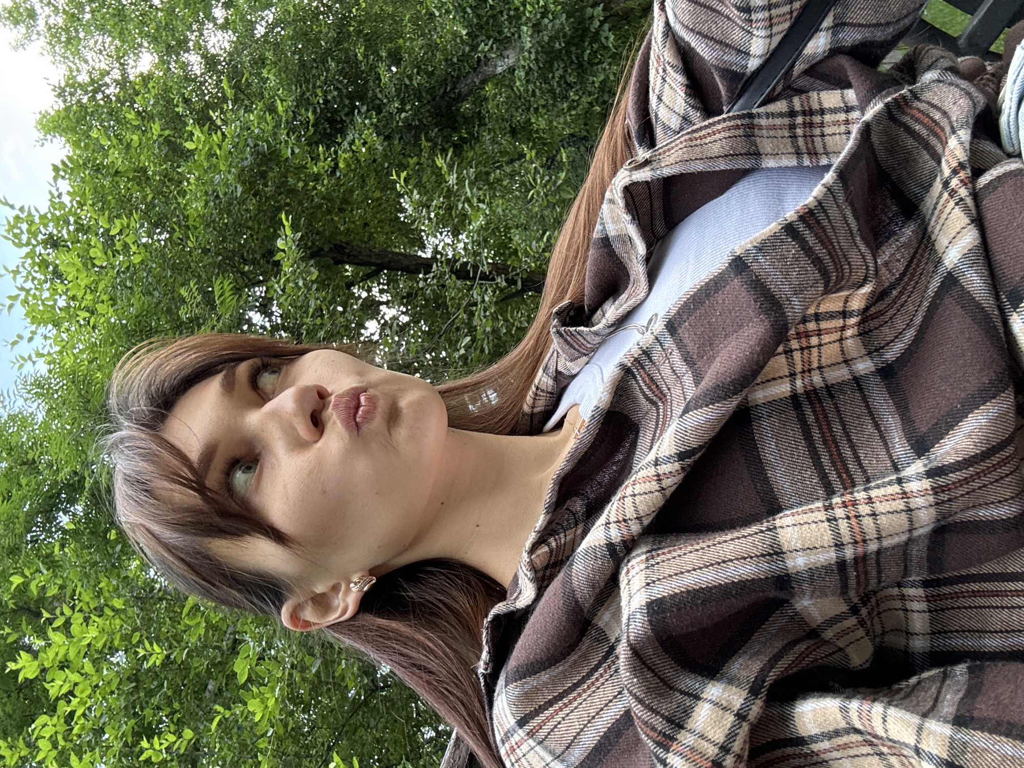 An outdoor medium shot shows a young woman sitting and posing for a selfie. She has long brown hair, light skin, and is wearing a brown and beige plaid shirt over a white top. Her lips are pursed as if she is blowing a kiss, and she is looking off to her left. The background consists of lush green trees and a blue sky, suggesting a park or a similar natural setting within the city of Novokuznetsk, Russia.