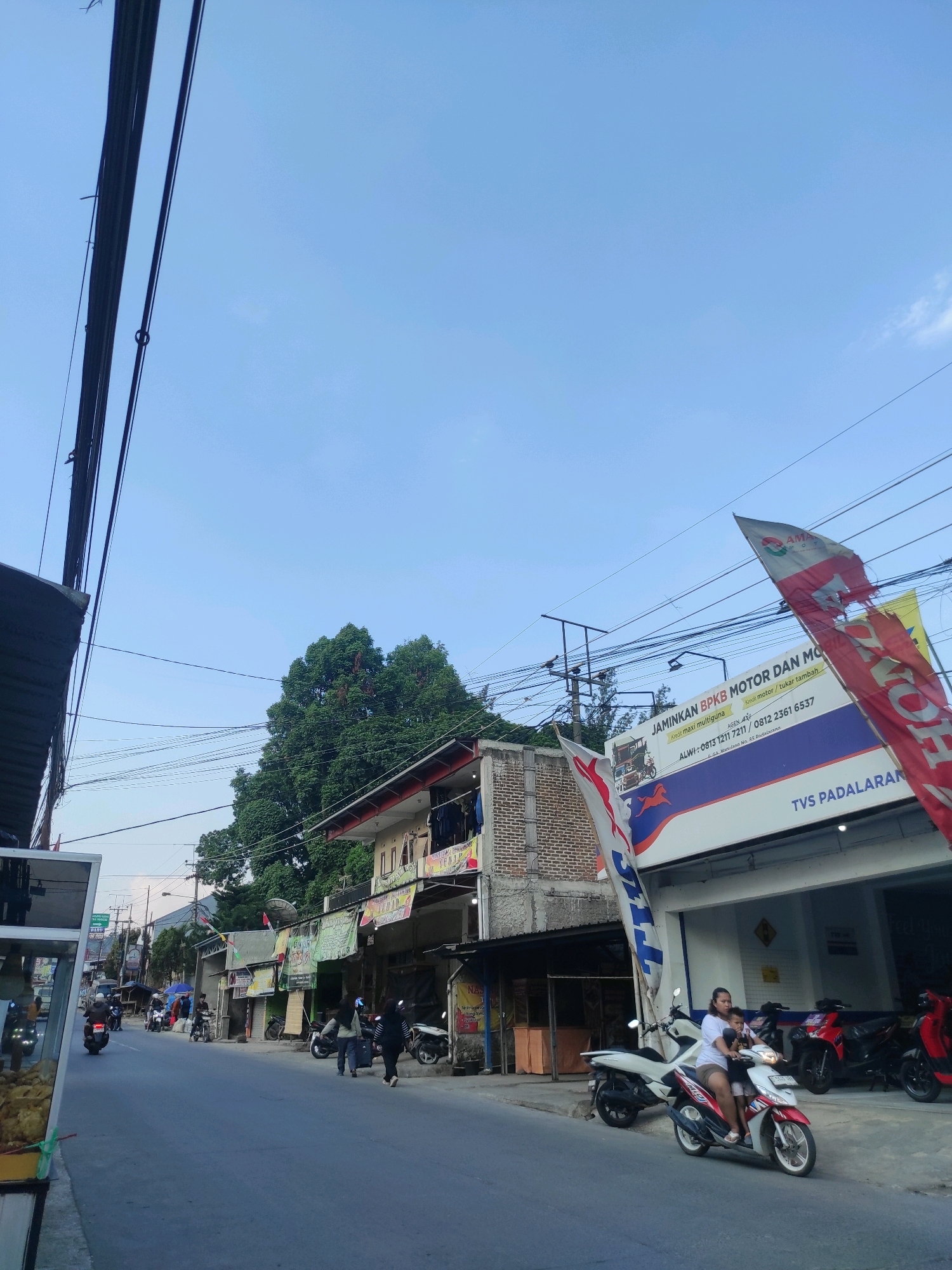 The image shows a street scene in Padalarang, Indonesia. The street is paved and appears to be a two-lane road. On the left side of the street, there are buildings with storefronts, and on the right side, there is a motorcycle dealership with several motorcycles displayed outside.
Several people are visible in the image, mostly walking or riding motorcycles. One woman is riding a white and red scooter with a child in front of her. There are also several parked motorcycles and scooters on the sidewalk.
The buildings along the street appear to be commercial establishments, with various signs and banners displayed. One prominent banner advertises "JAMINIKAN BPKB MOTOR DAN MO". The sky is clear and blue, with a few wispy clouds. There are also many overhead power lines visible, a common sight in urban areas of Indonesia. The overall impression is that of a busy street in a commercial district.