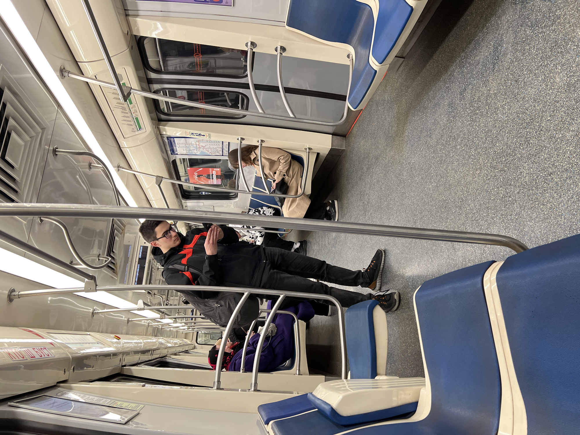 Inside a subway car in Saint Petersburg, Russia, a man lies across multiple seats. The interior of the subway car features white walls, blue and white seats, and metal handrails. The floor is a gray speckled material. Several passengers are present in the car. The man laying across the seats is wearing a black jacket with red accents, black pants, and sneakers. He has dark hair, wears glasses, and is looking towards the right of the frame. Other passengers are seated nearby, including a woman in a beige coat looking at her phone, and another person in a purple jacket. Subway maps and informational signs are visible on the walls of the subway car.